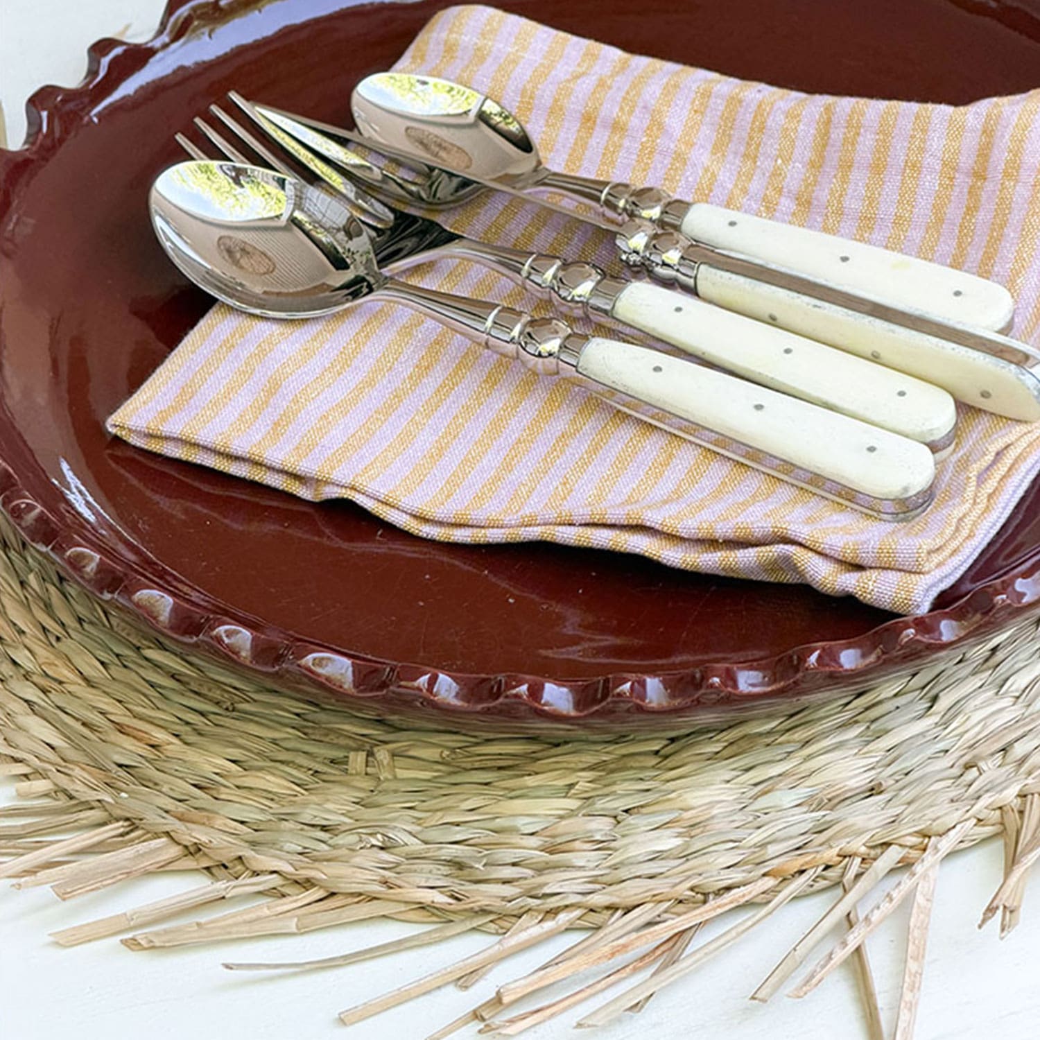 Juego de cubiertos con mangos blancos sobre servilleta a rayas en plato marrón y base trenzada