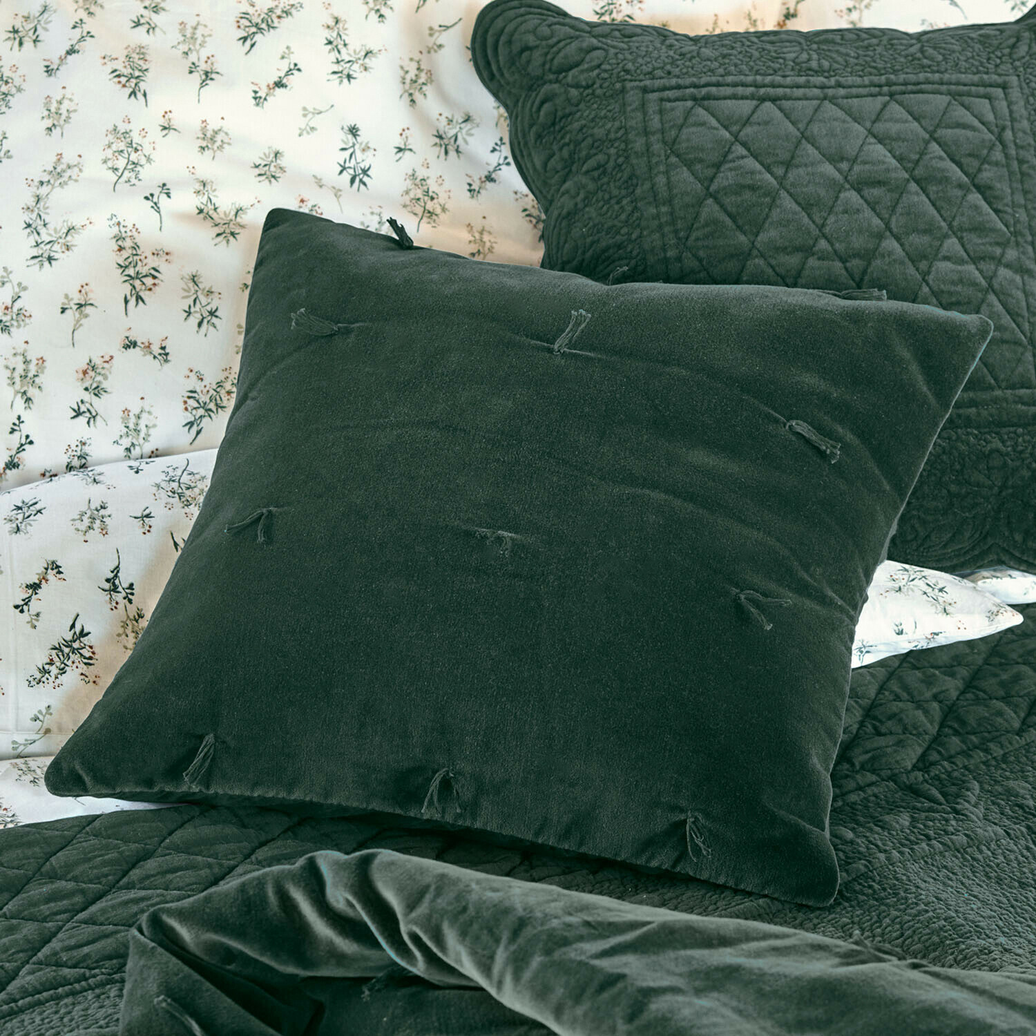 Cojín Orélisse Cojín decorativo verde de terciopelo con borlas sobre cama con colcha acolchada y ropa de cama floral