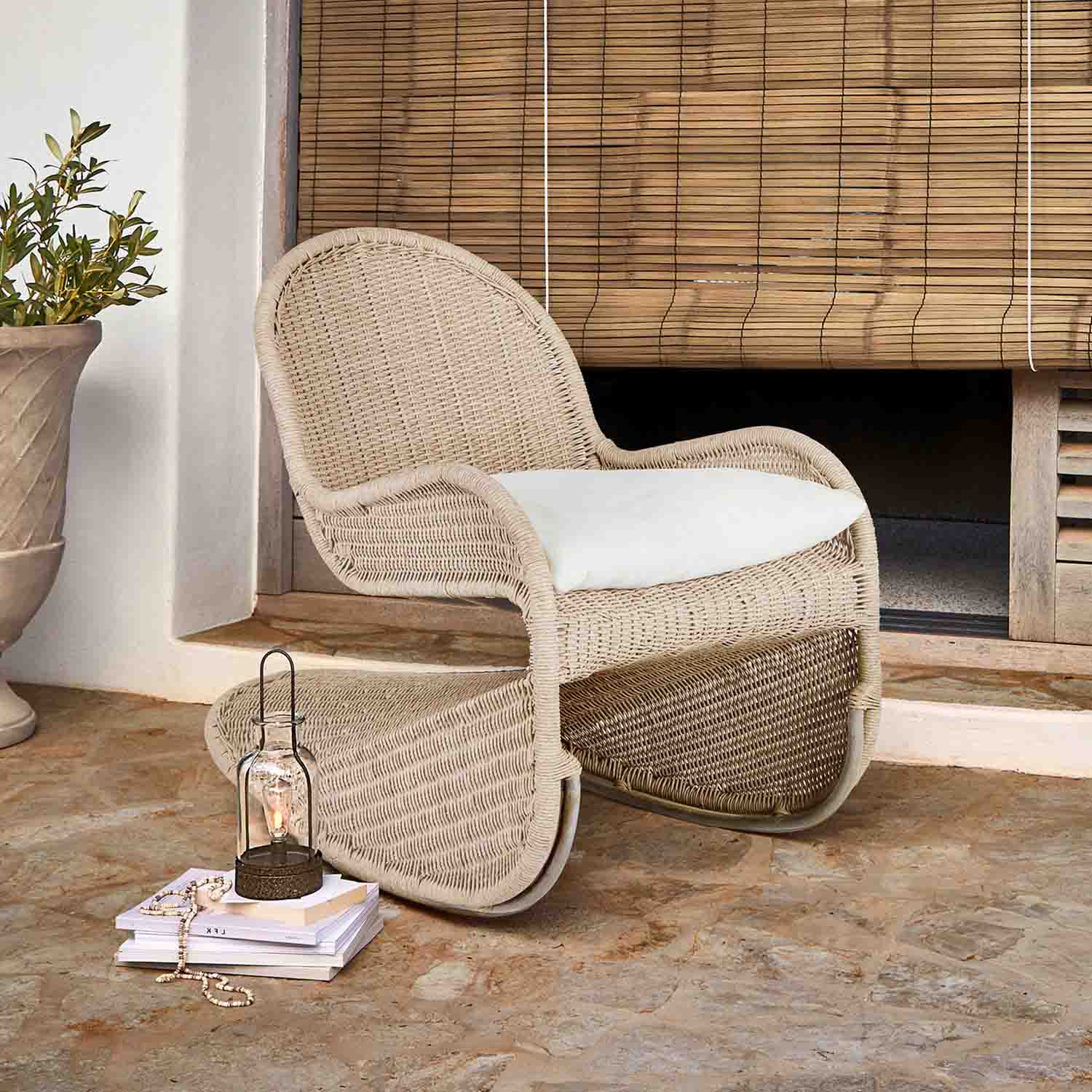 Sillón de mimbre beige con cojín blanco en terraza, junto a farol y libros sobre suelo de piedra