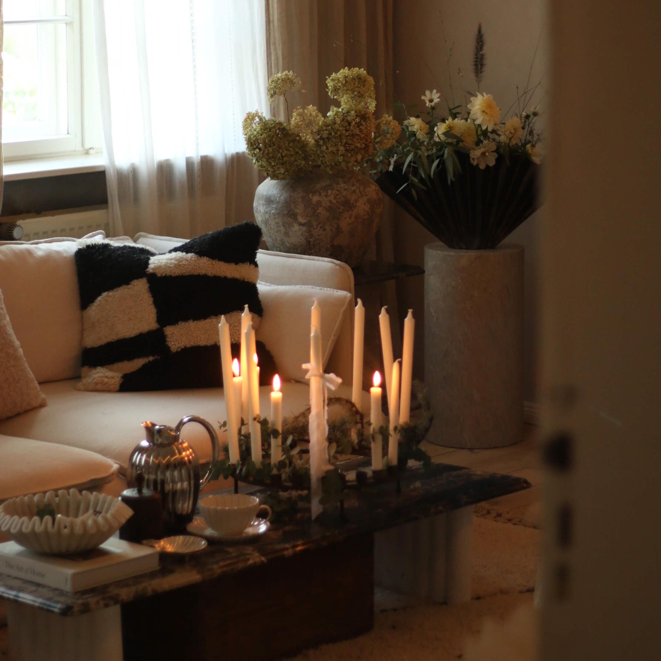 Sala acogedora con sofá crema, cojín blanco y negro y velas encendidas sobre mesa de mármol