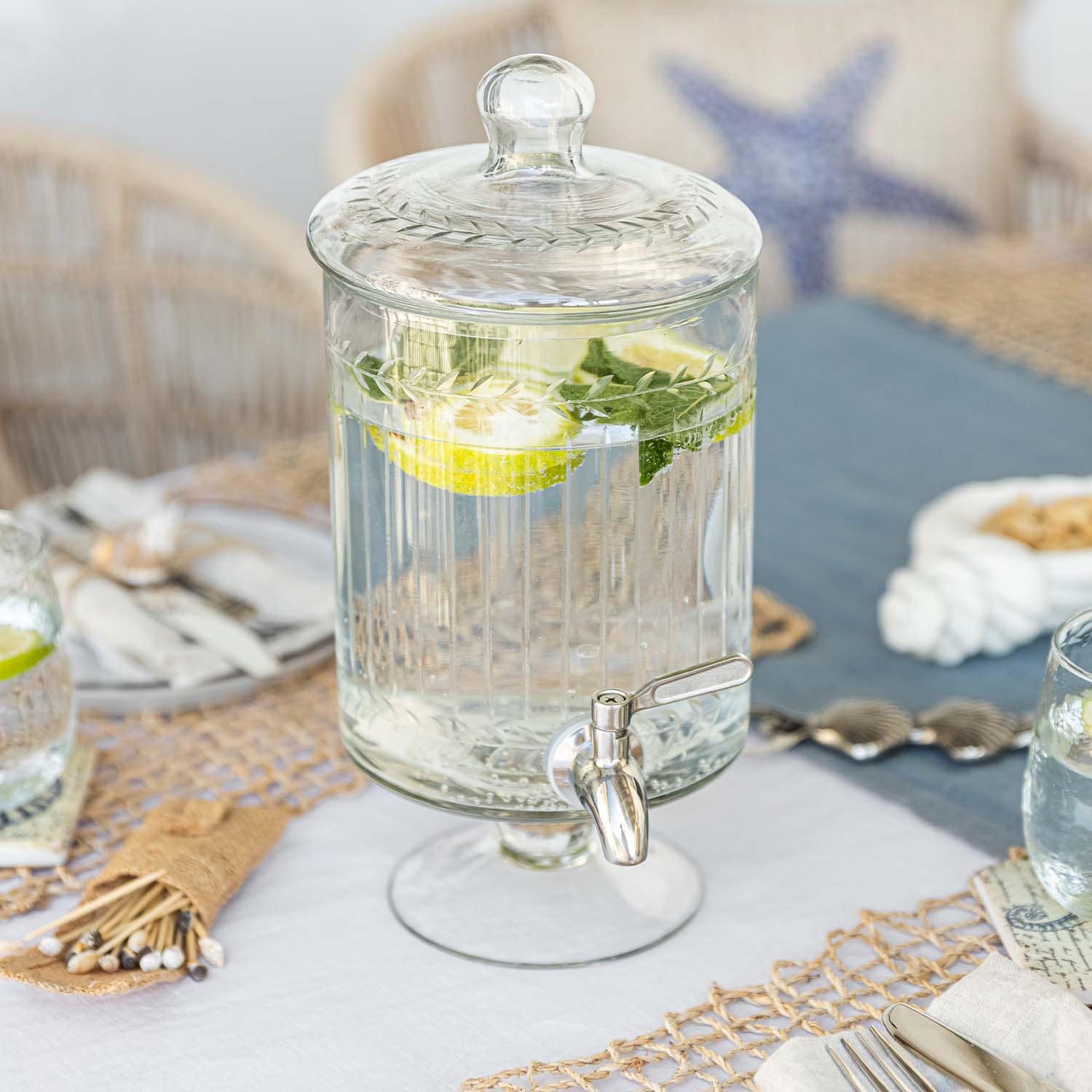 Dispensador de bebidas de vidrio con tapa y grifo, agua con limón y menta sobre mesa