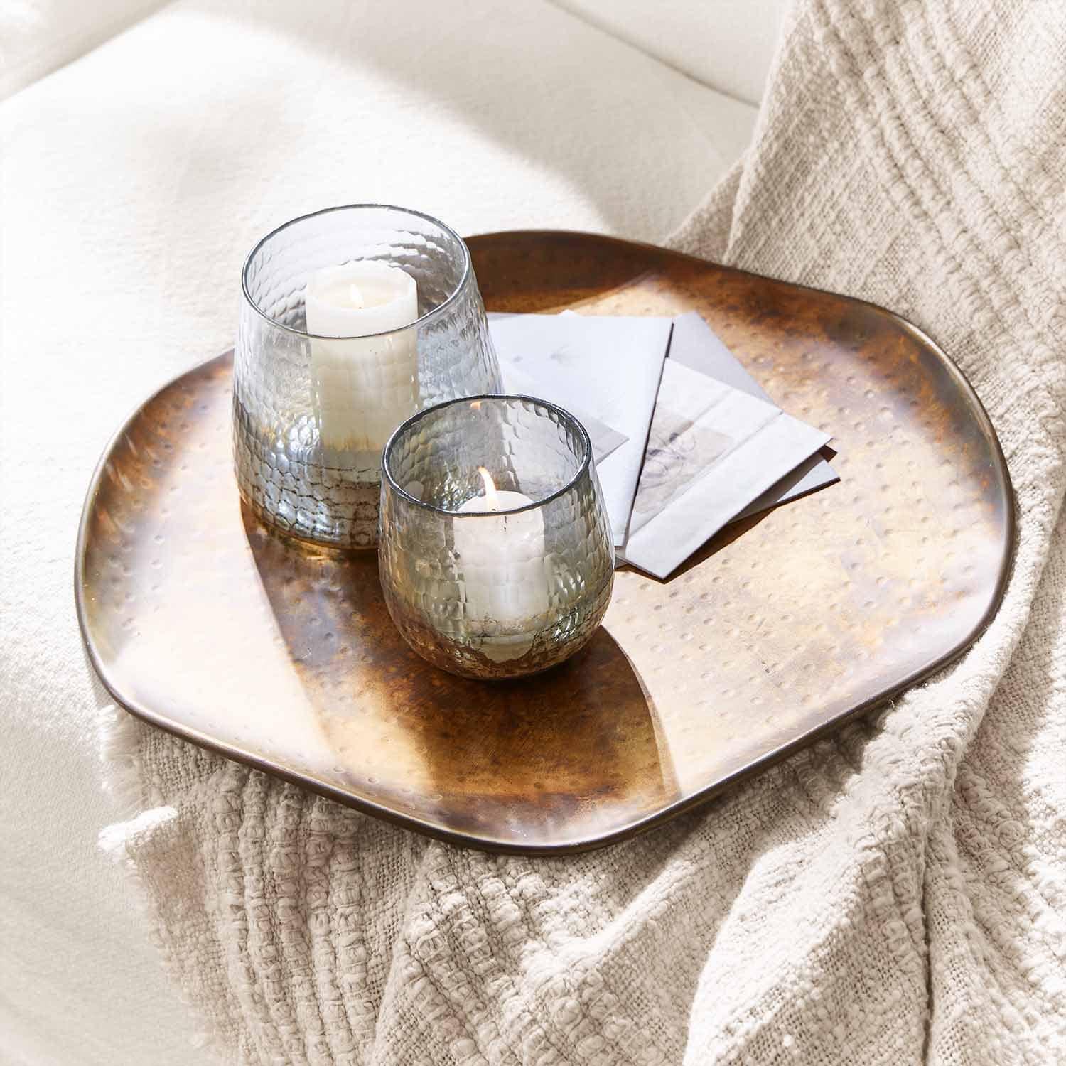 Dos portavelas de vidrio con velas blancas sobre una bandeja de cobre sobre una manta clara para decoración