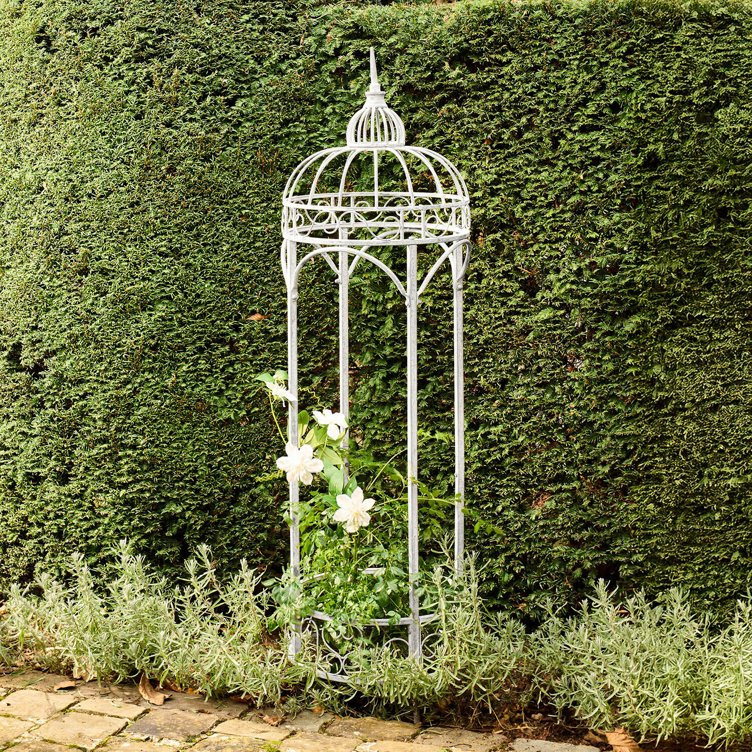 Templete de jardín de metal blanco con diseño decorativo, sosteniendo plantas trepadoras frente a un seto verde