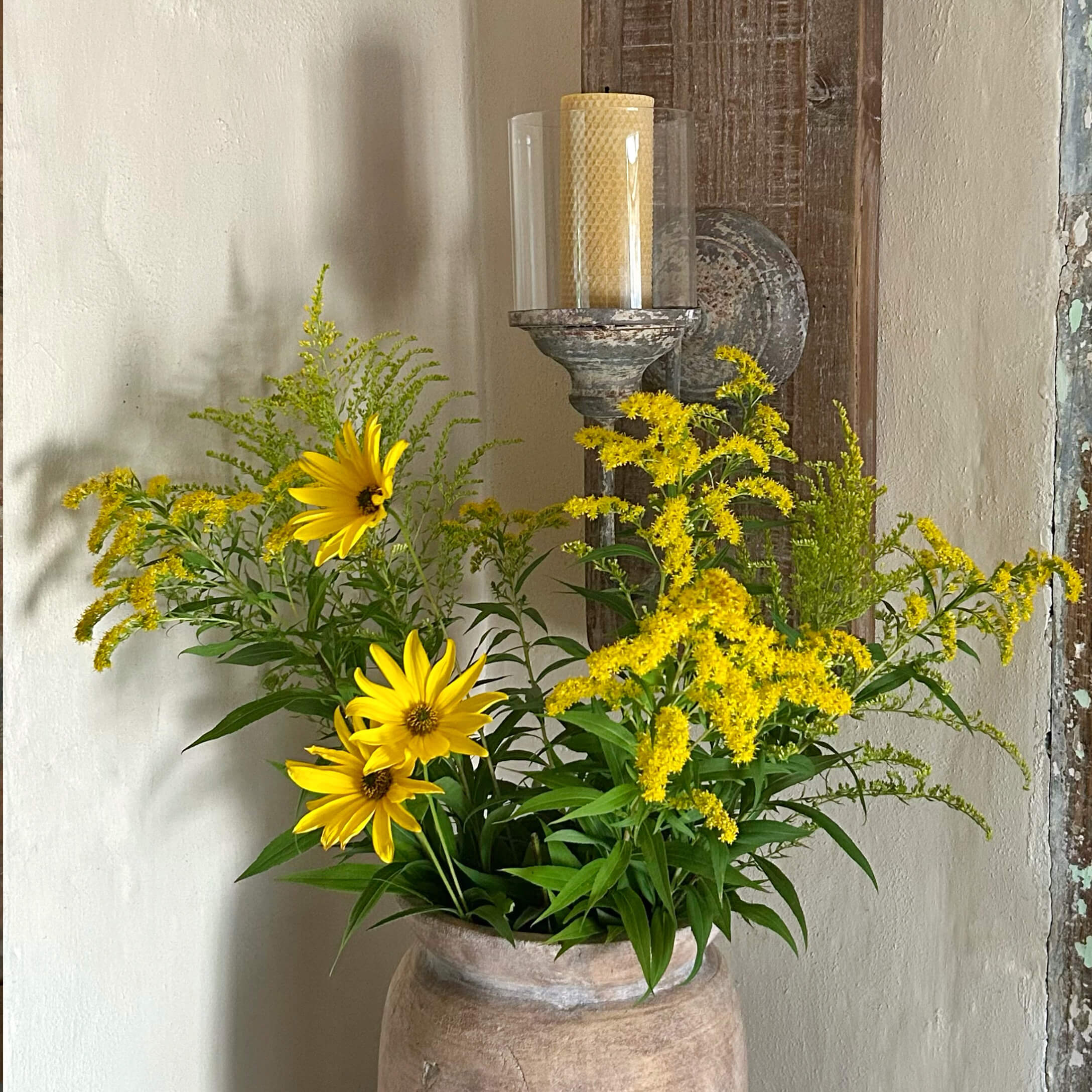 Flores amarillas en un jarrón de barro frente a una pared con un candelabro rústico