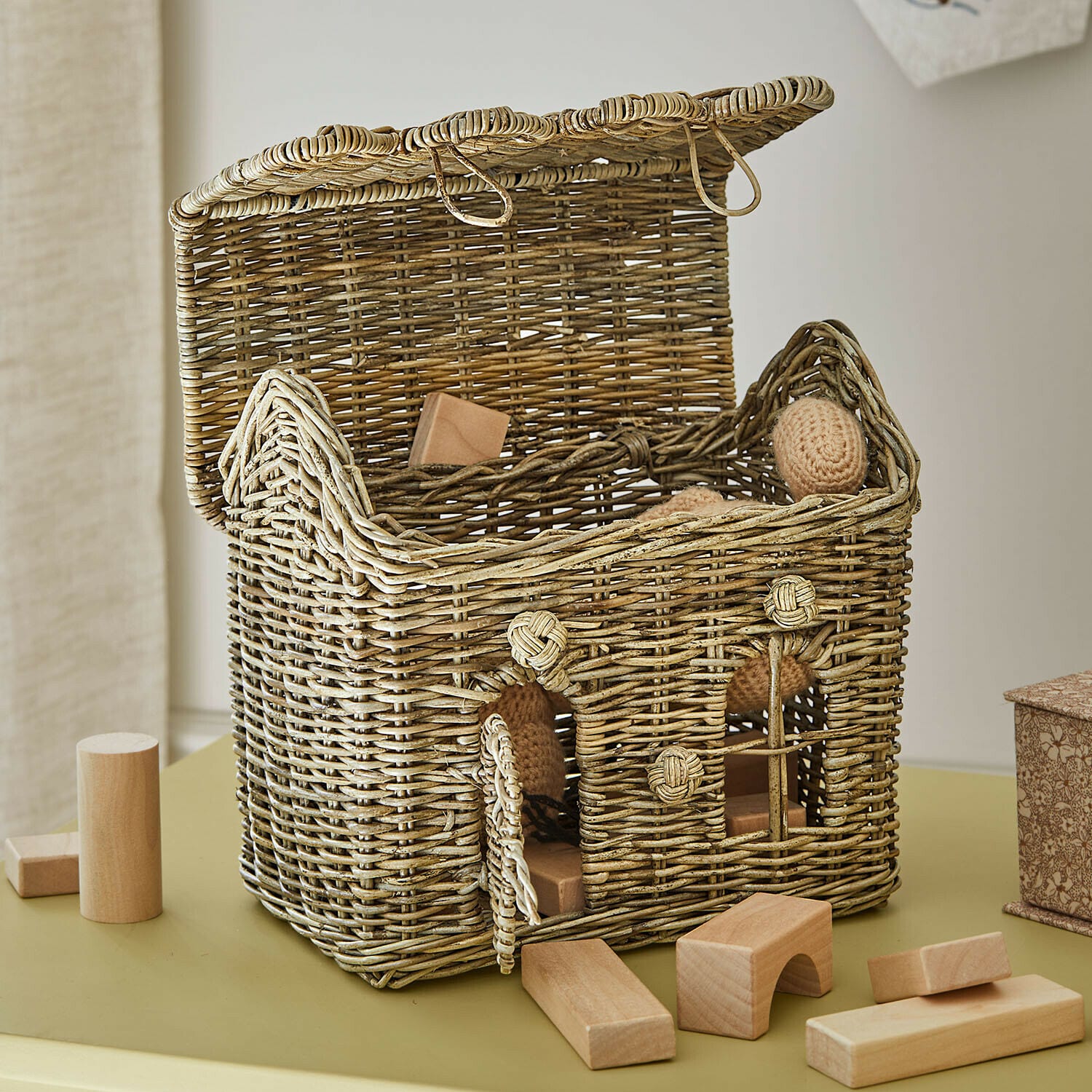 Cesta de mimbre abierta en forma de casa con bloques de madera y juguetes en colores naturales