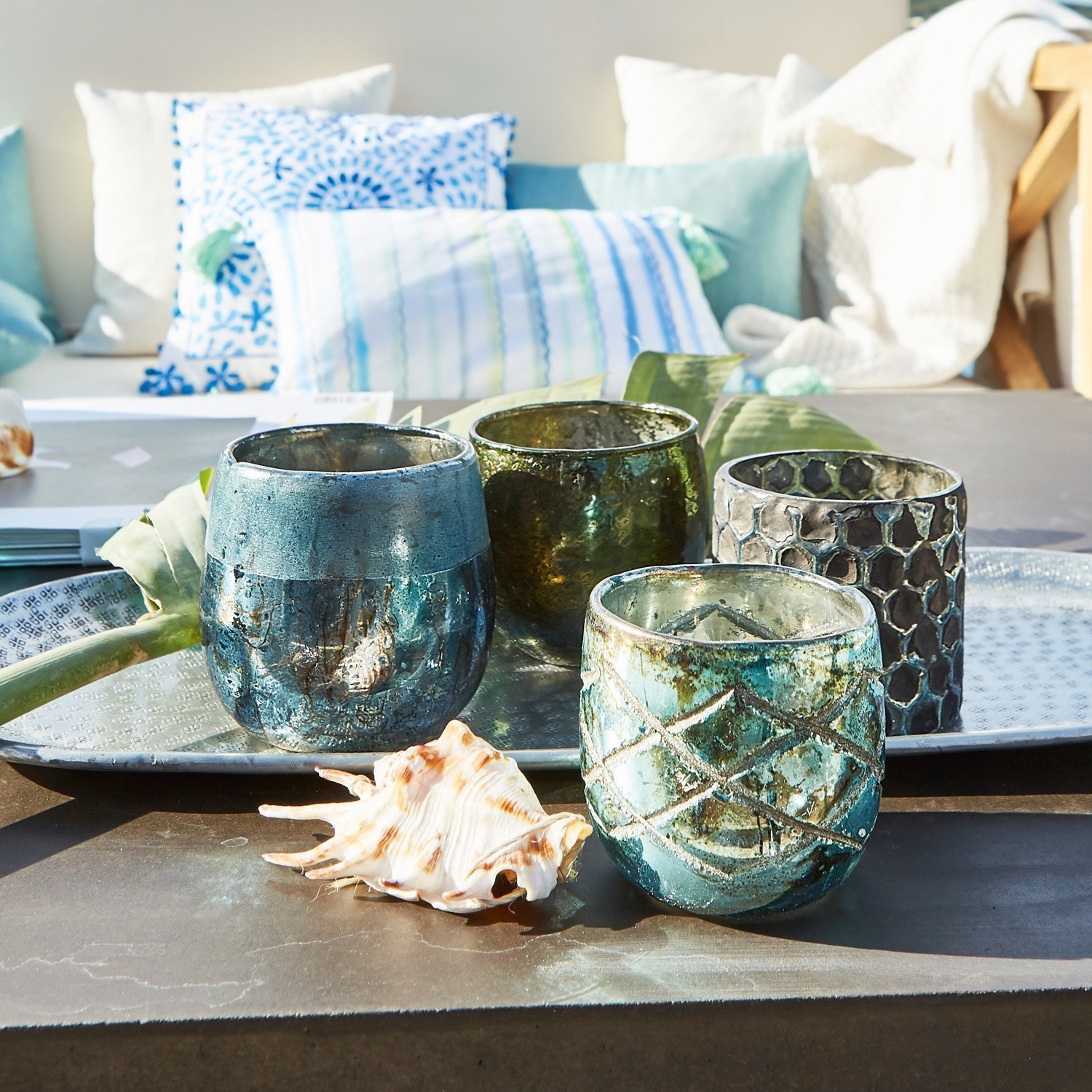 Conjunto de cuatro portavelas de vidrio en tonos azules y verdes sobre bandeja metálica con concha decorativa