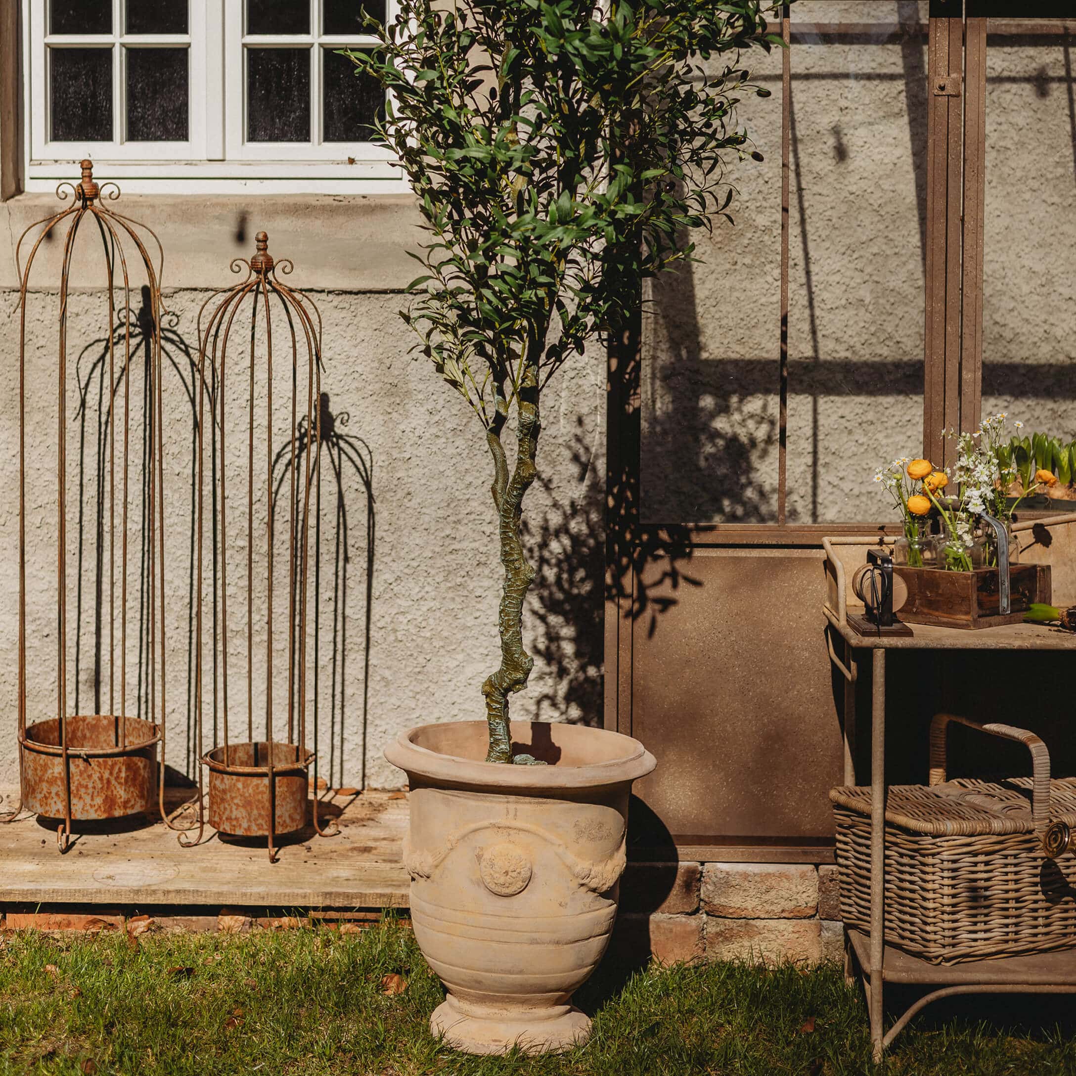 Maceta grande de cerámica beige con árbol verde en jardín con decoraciones metálicas oxidadas