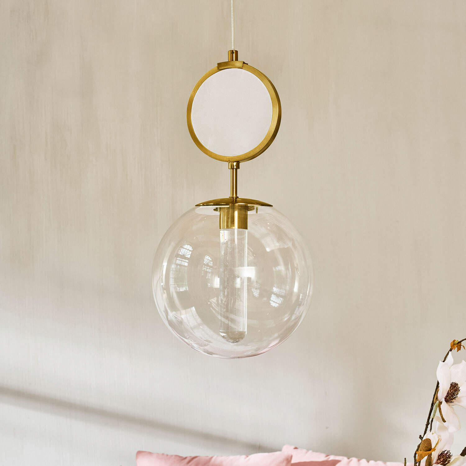 Lámpara colgante dorada con esfera de vidrio transparente y elemento decorativo circular sobre pared clara