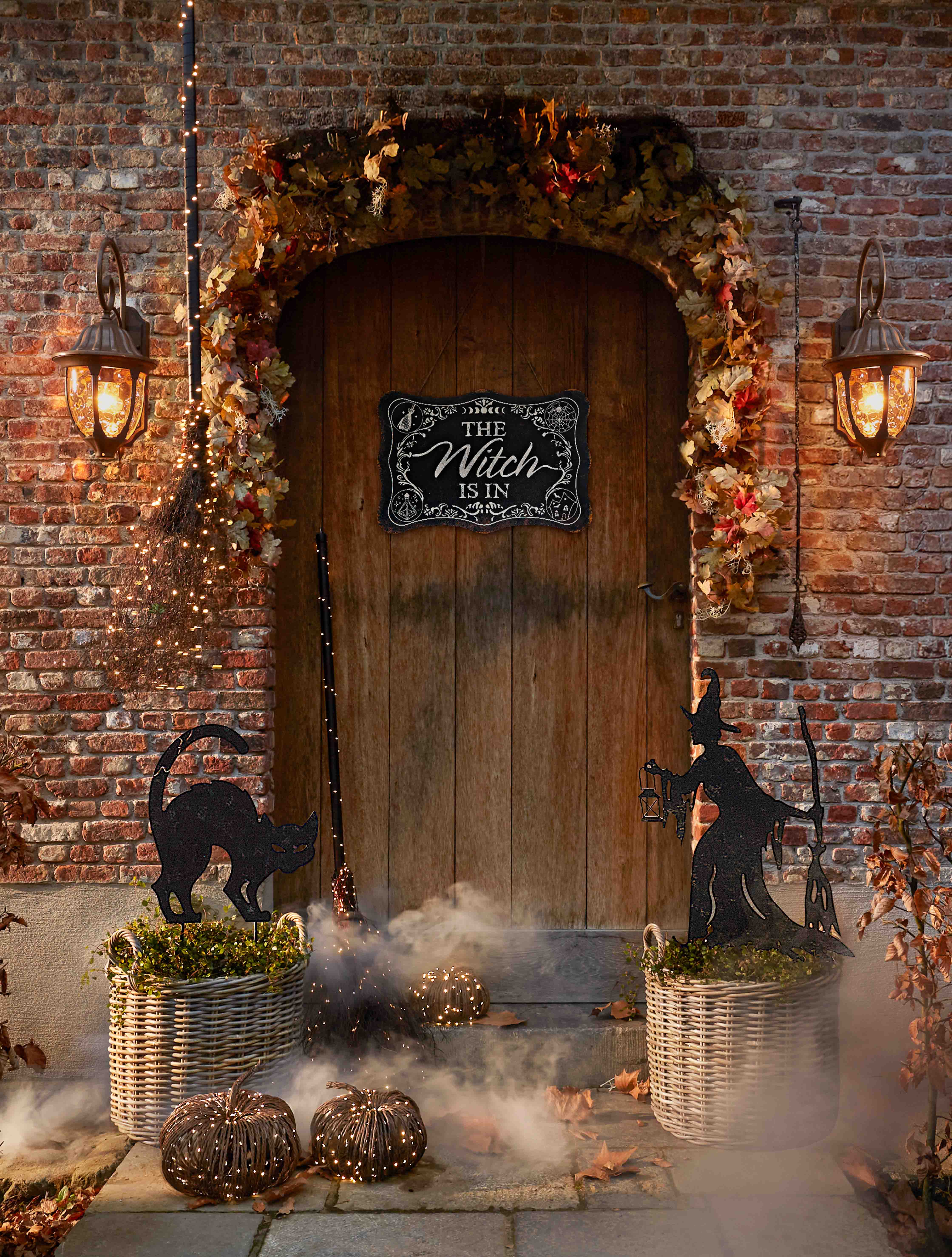Decoración de Halloween con bruja, gato negro y faroles frente a puerta de madera y pared de ladrillo