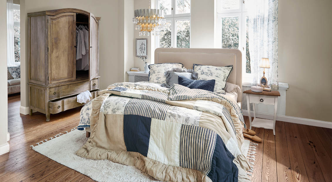 Dormitorio con colcha patchwork en beige, azul marino y blanco sobre cama con cabecero tapizado