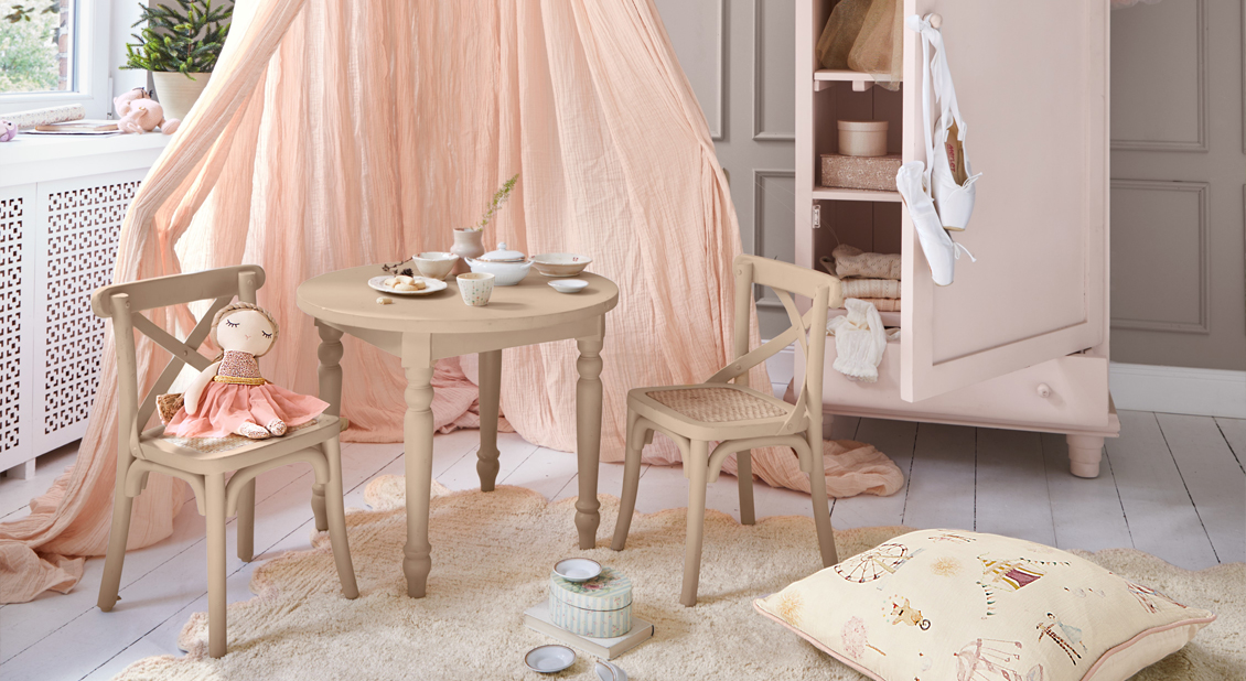Mesa y sillas infantiles de madera beige con muñeca y cortina rosa empolvado en habitación infantil pastel