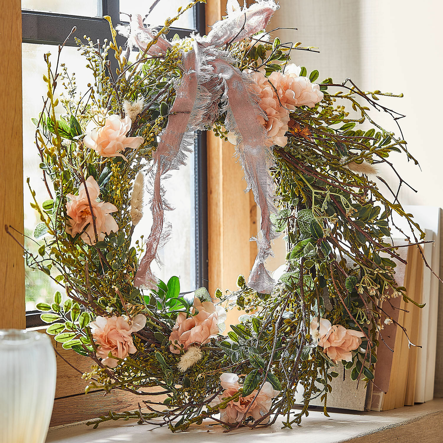 Corona Chérisse Corona decorativa de ramas, flores rosas y hojas verdes con lazo rosa en la ventana