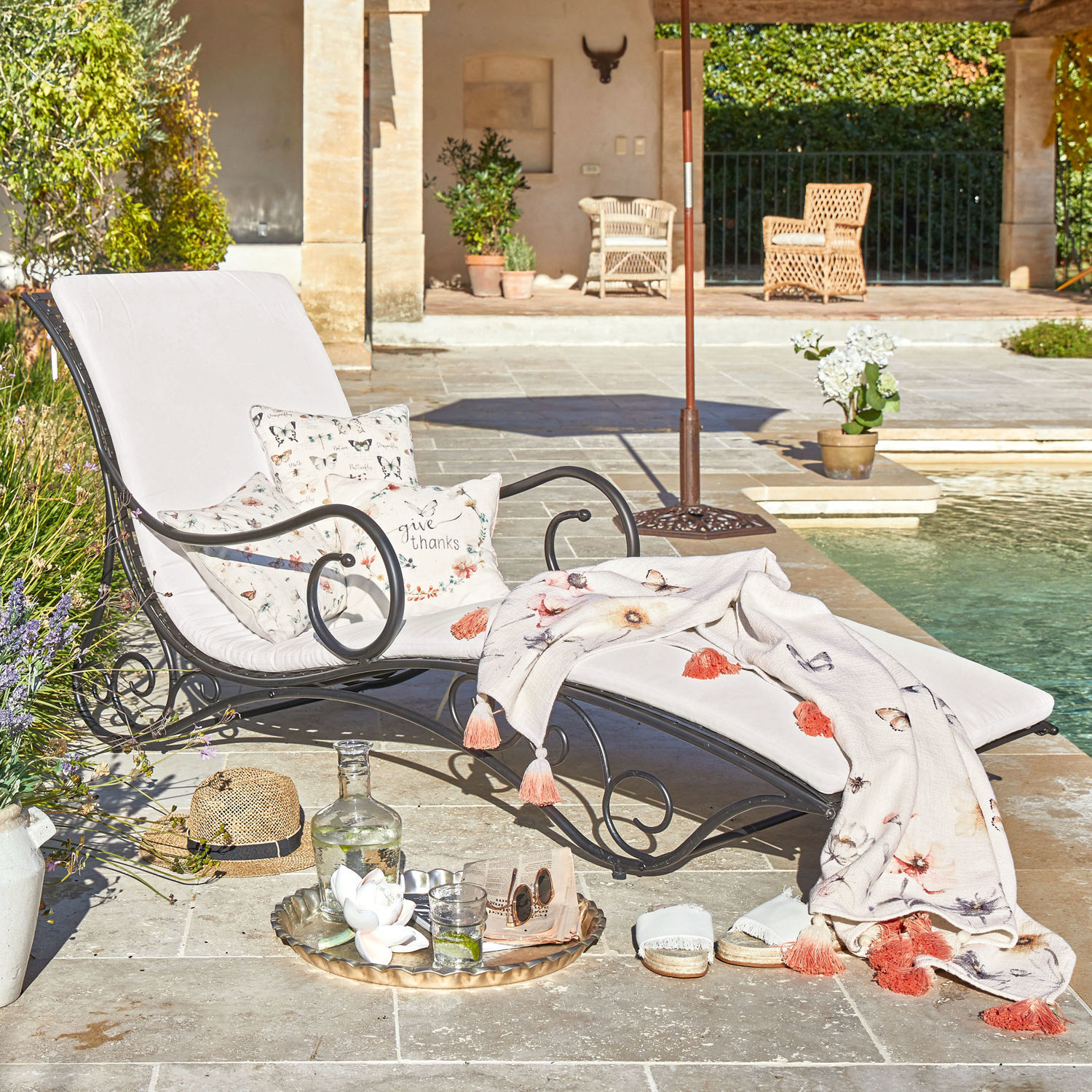 Tumbona de jardín con estructura metálica negra y cojines claros, decorada con manta y cojines con motivos florales y mariposas junto a la piscina