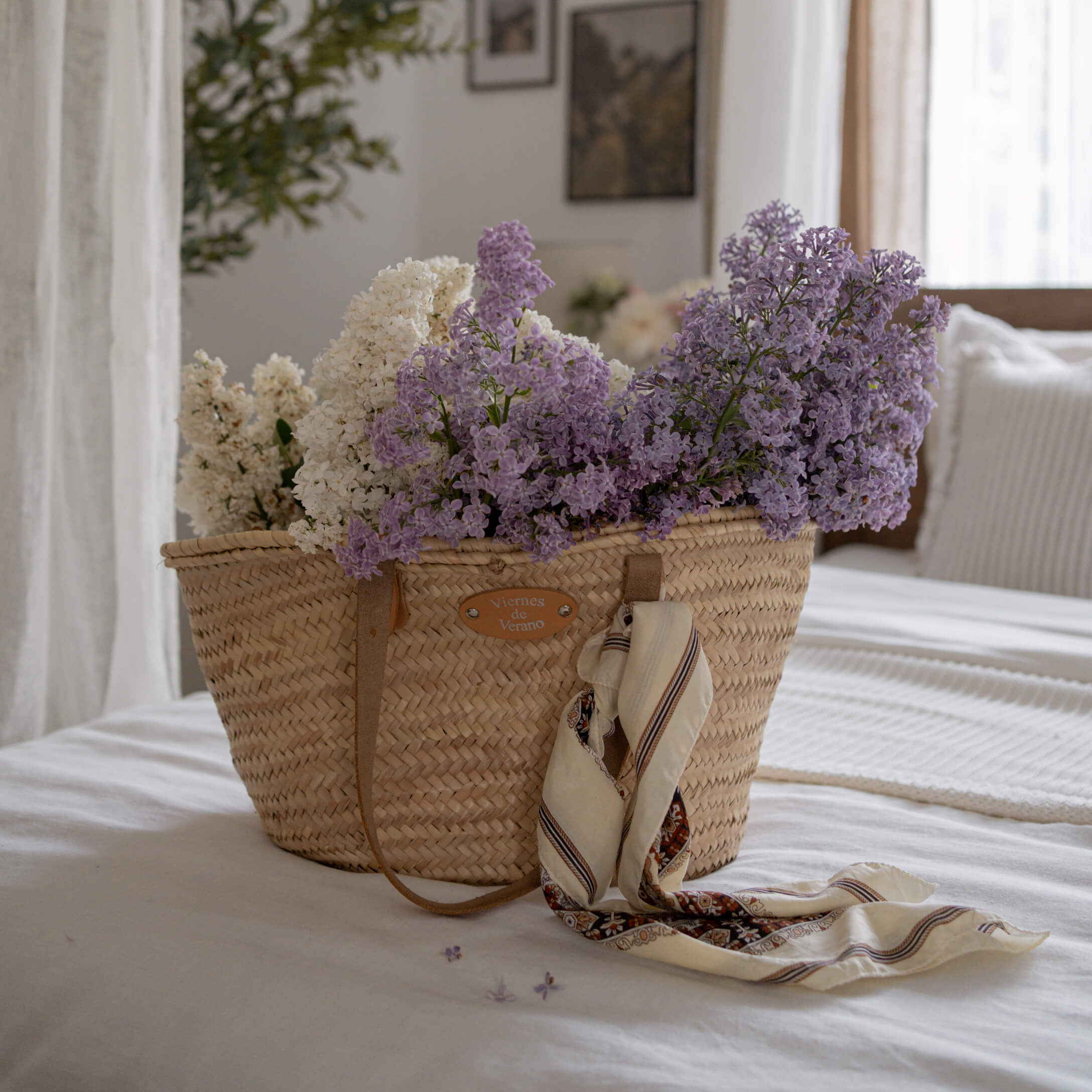 Cesta de mimbre con flores blancas y moradas y pañuelo sobre cama blanca en habitación luminosa