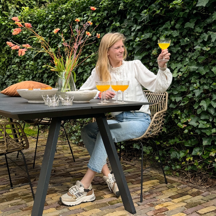 Mujer con blusa blanca y vaqueros sentada en mesa negra al aire libre, sostiene copa con bebida naranja, jarrón con flores al lado