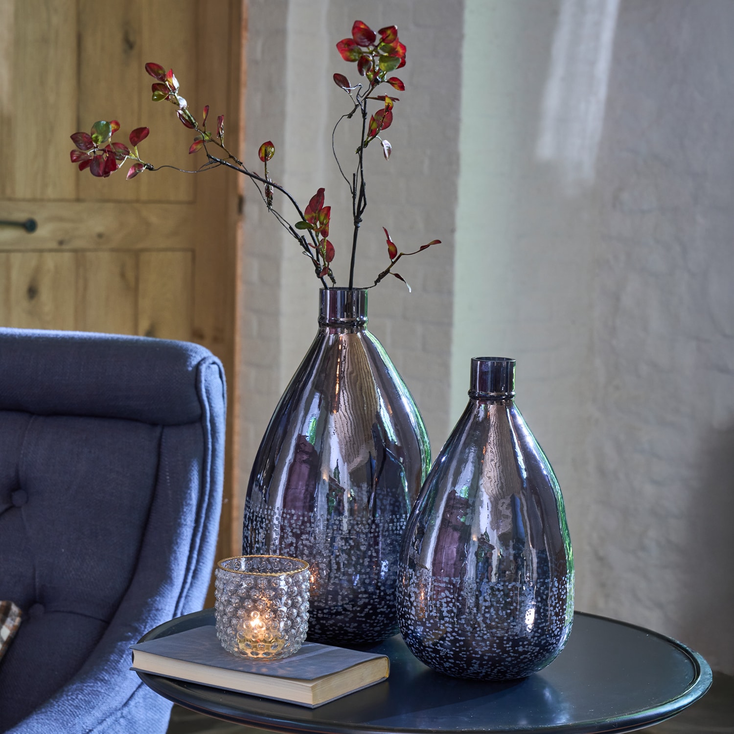 Dos jarrones decorativos de vidrio gris oscuro con ramas rojas sobre una mesa junto a un portavelas y un libro