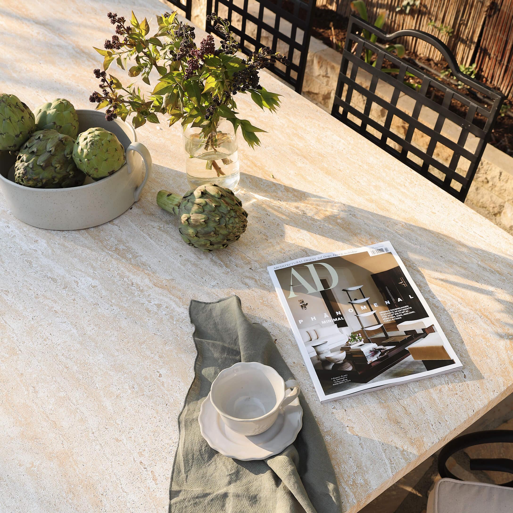 Mesa de piedra clara con alcachofas verdes, taza blanca y revista de decoración al aire libre