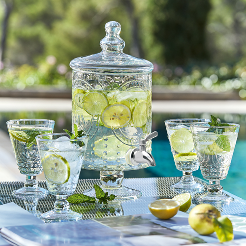Dispensador de bebidas de vidrio con limón y menta, cuatro vasos sobre mesa exterior junto a piscina