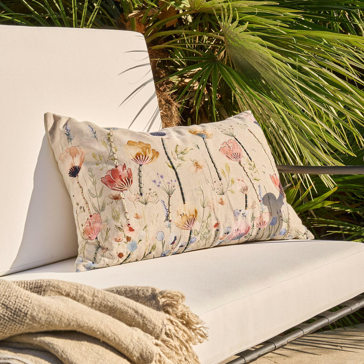 Funda de cojín Cadrinela Cojín beige con diseño floral sobre sillón exterior claro con manta beige