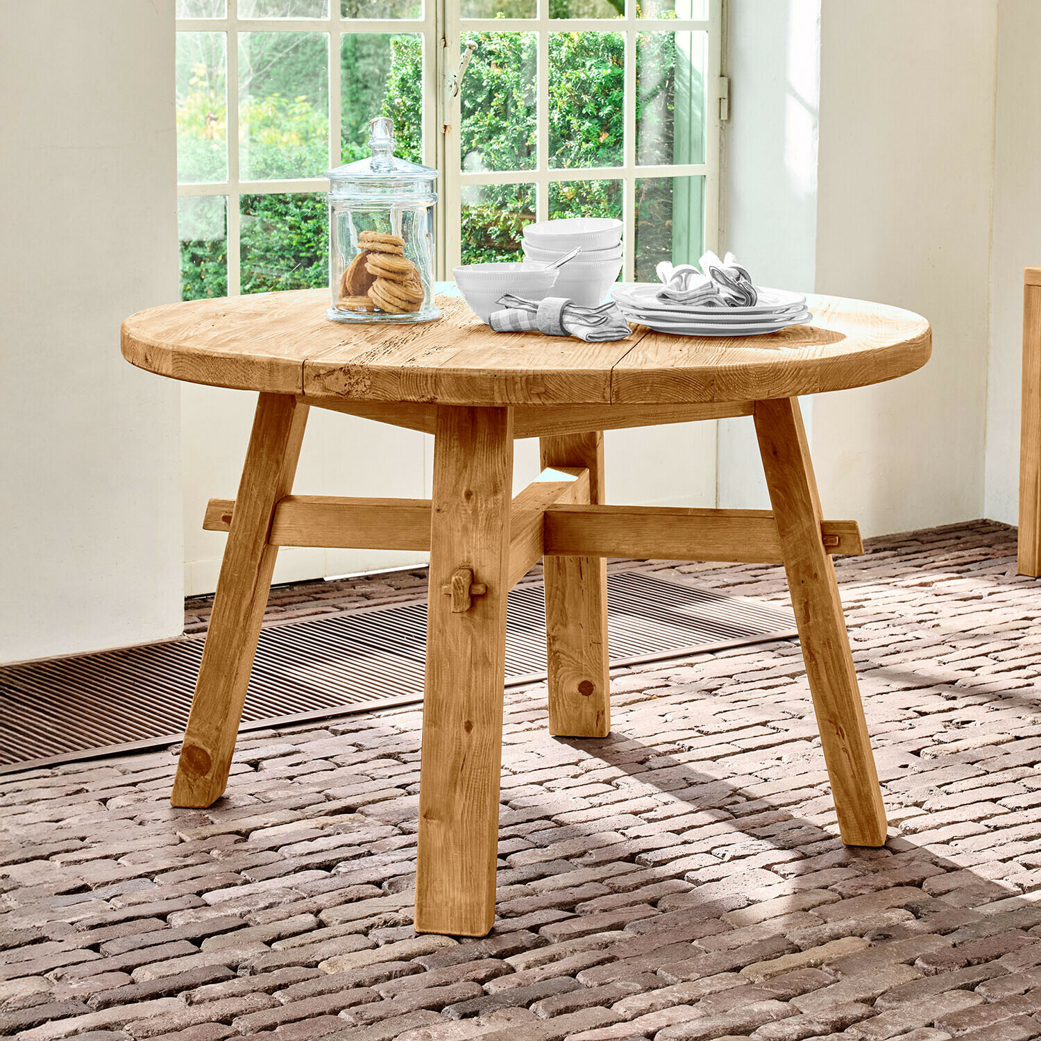 Mesa redonda de madera de pino natural sobre suelo de ladrillo junto a ventana luminosa