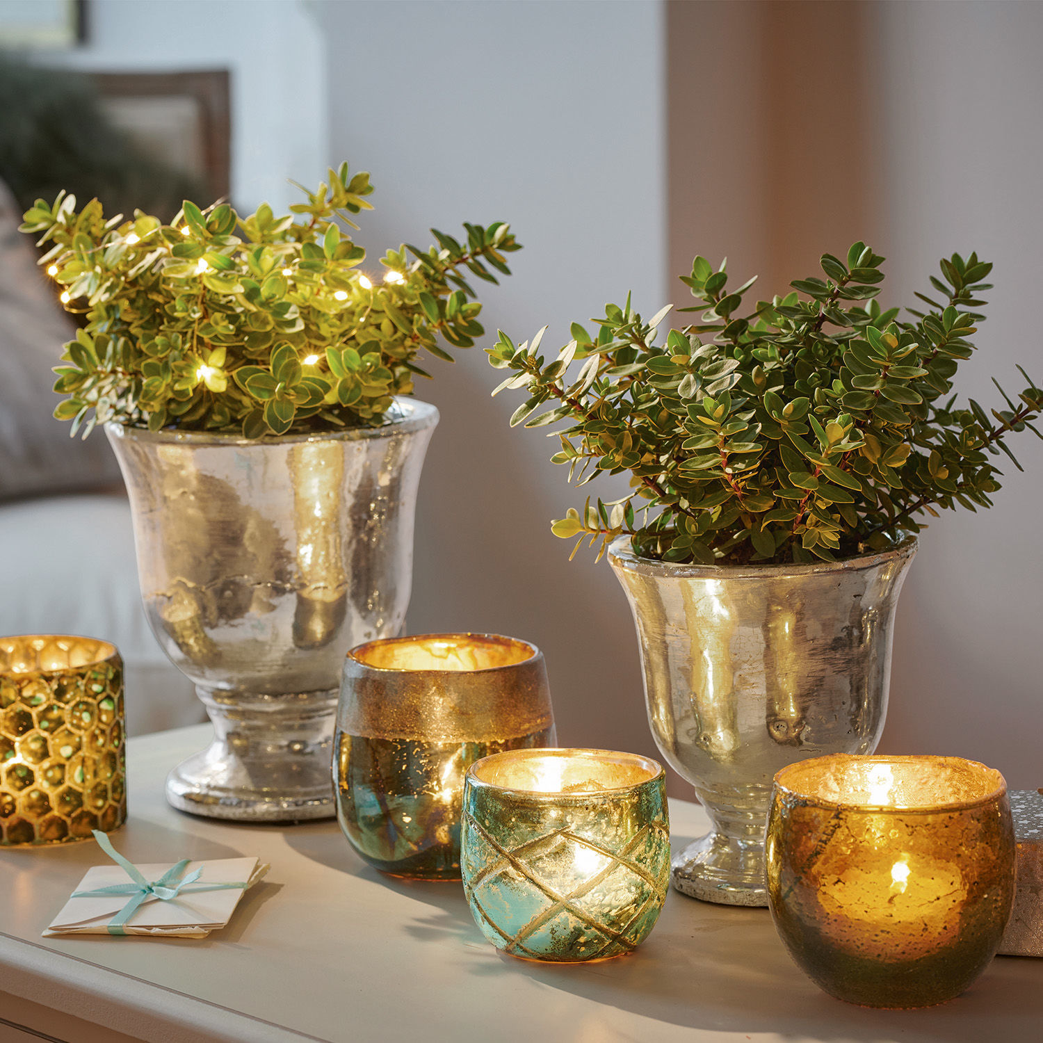 Macetas decorativas plateadas con plantas verdes y portavelas de vidrio con luz cálida sobre mesa