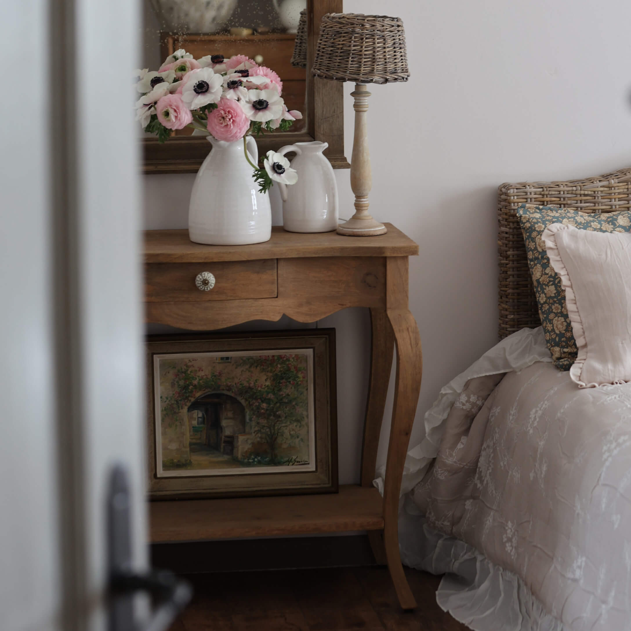 Mesa de madera con jarras de cerámica blanca y flores junto a cama con cabecero de mimbre