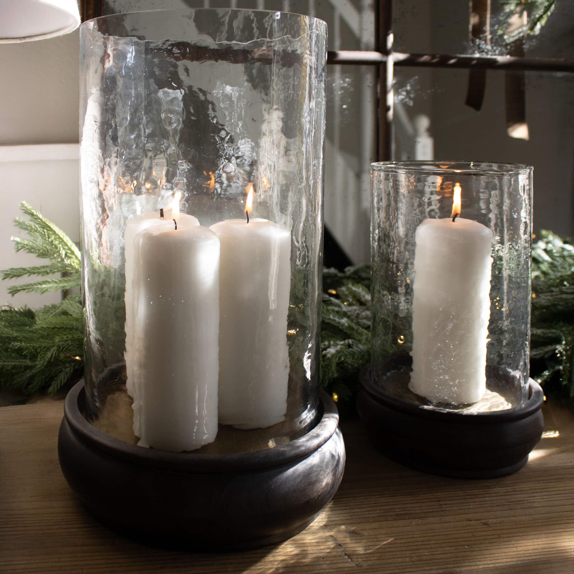 Faroles de vidrio con velas blancas sobre base de madera, decoración navideña con ramas verdes