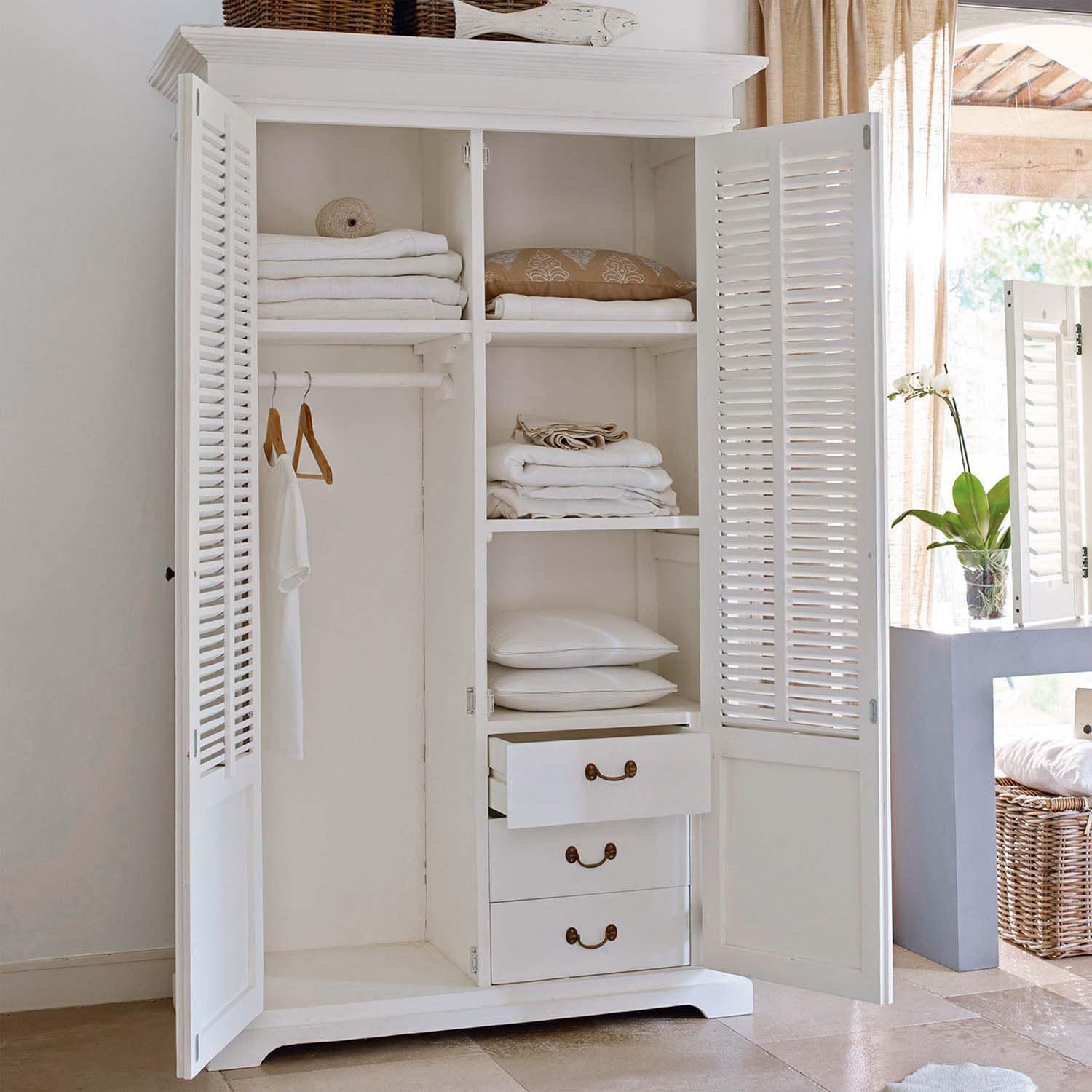 Armario de madera blanco con puertas abiertas, textiles blancos doblados y cojines en habitación luminosa