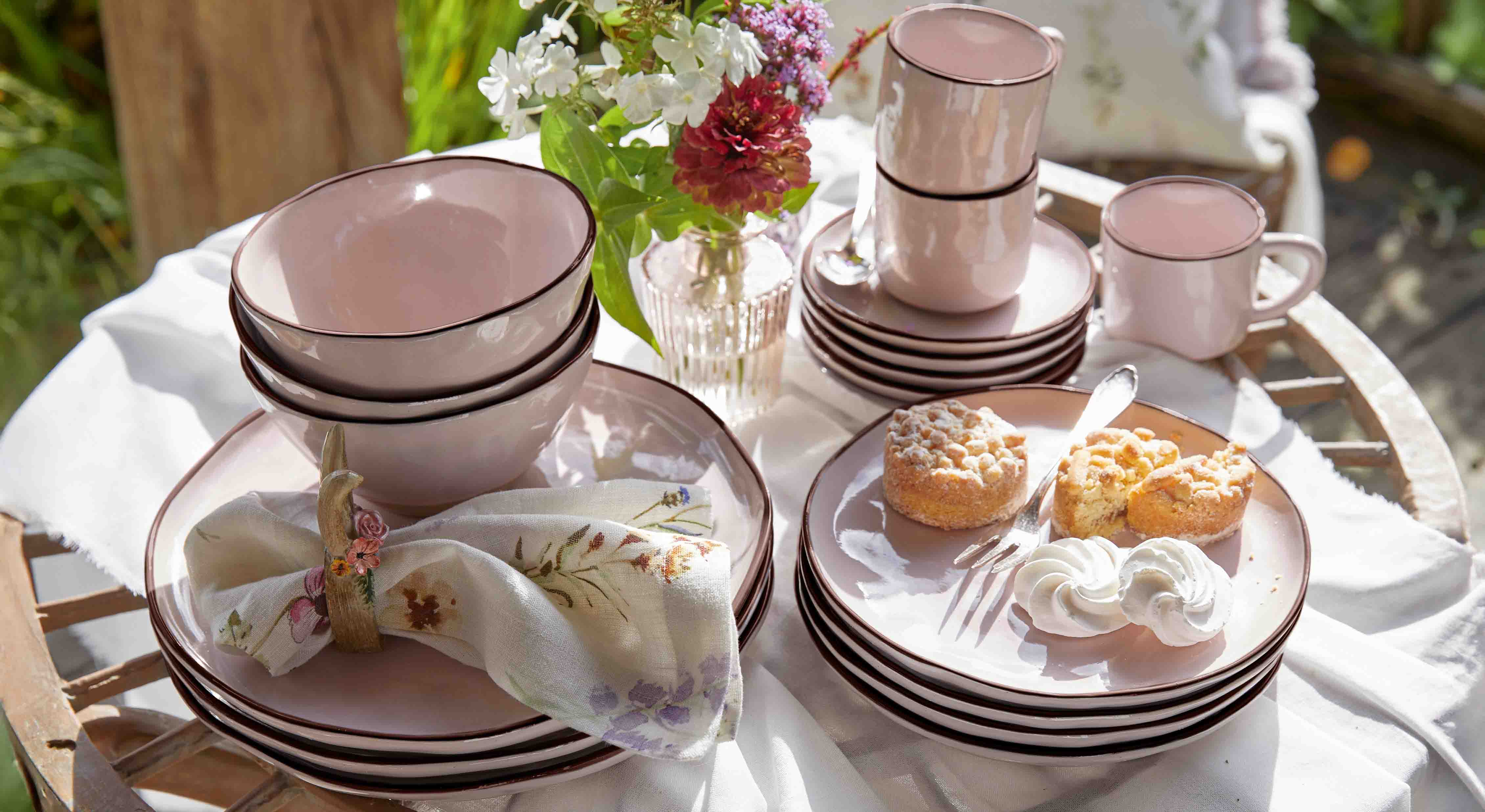 Juego de vajilla de cerámica rosa con platos, cuencos y tazas sobre mesa de madera con pasteles y flores