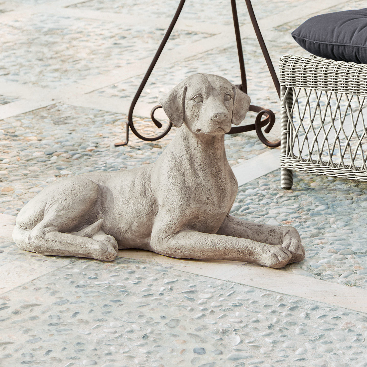 Perro Socrate Escultura de perro gris de hormigón tumbada sobre suelo de piedra junto a mueble de mimbre