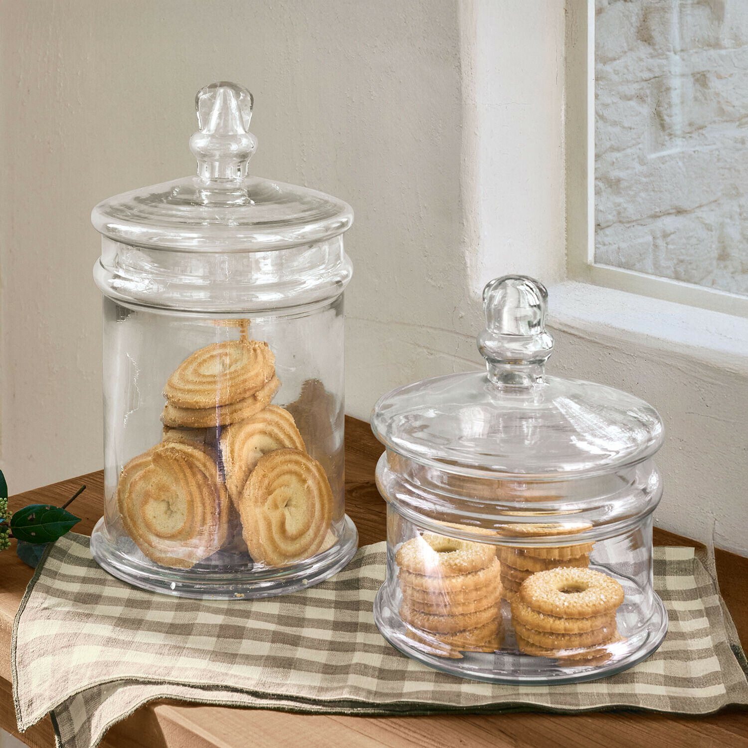 Dos recipientes de vidrio transparentes con tapas llenos de galletas sobre mesa de madera con mantel a cuadros