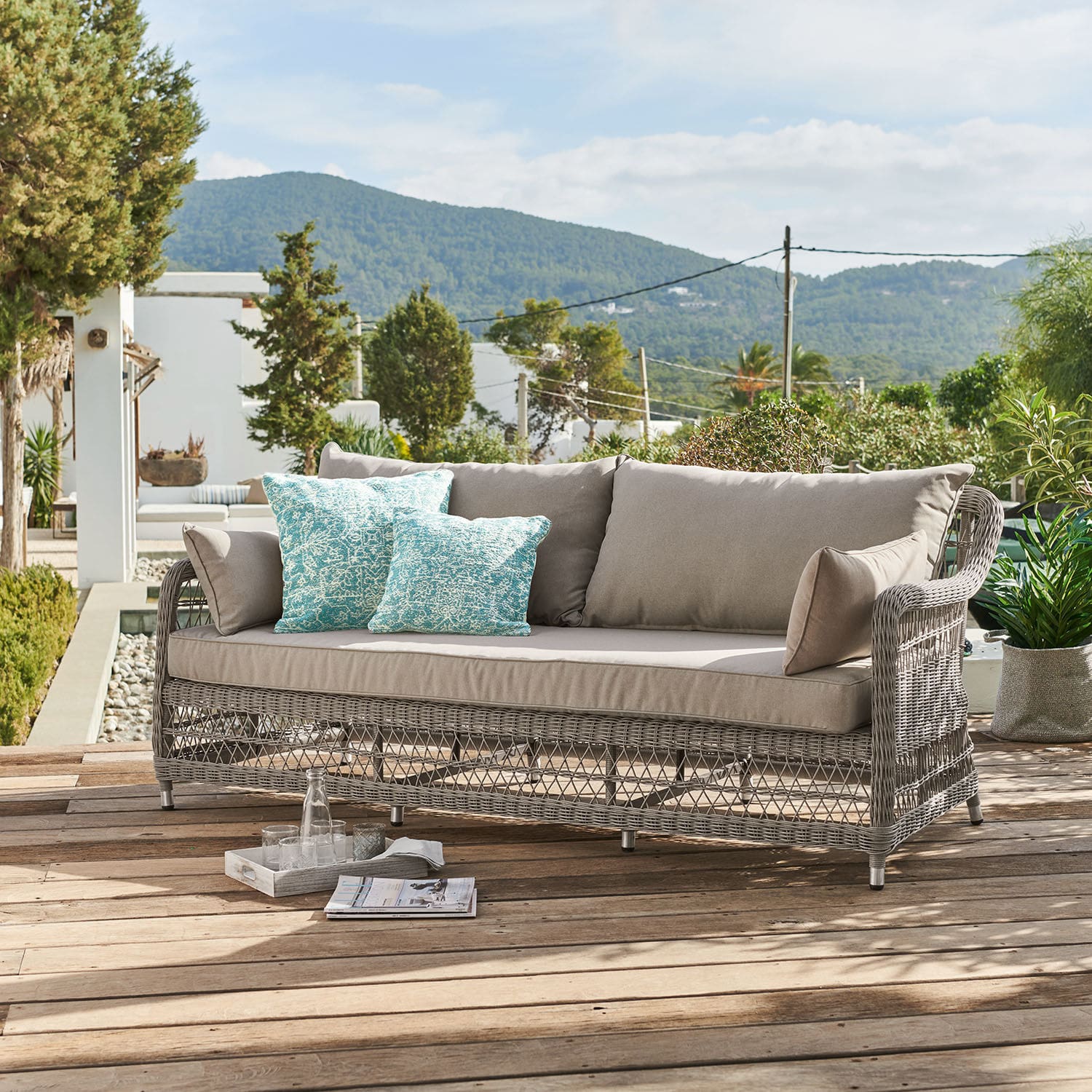 Sofá Tanaiste Sofá de jardín de ratán gris con cojines beige y cojines decorativos turquesa en terraza de madera