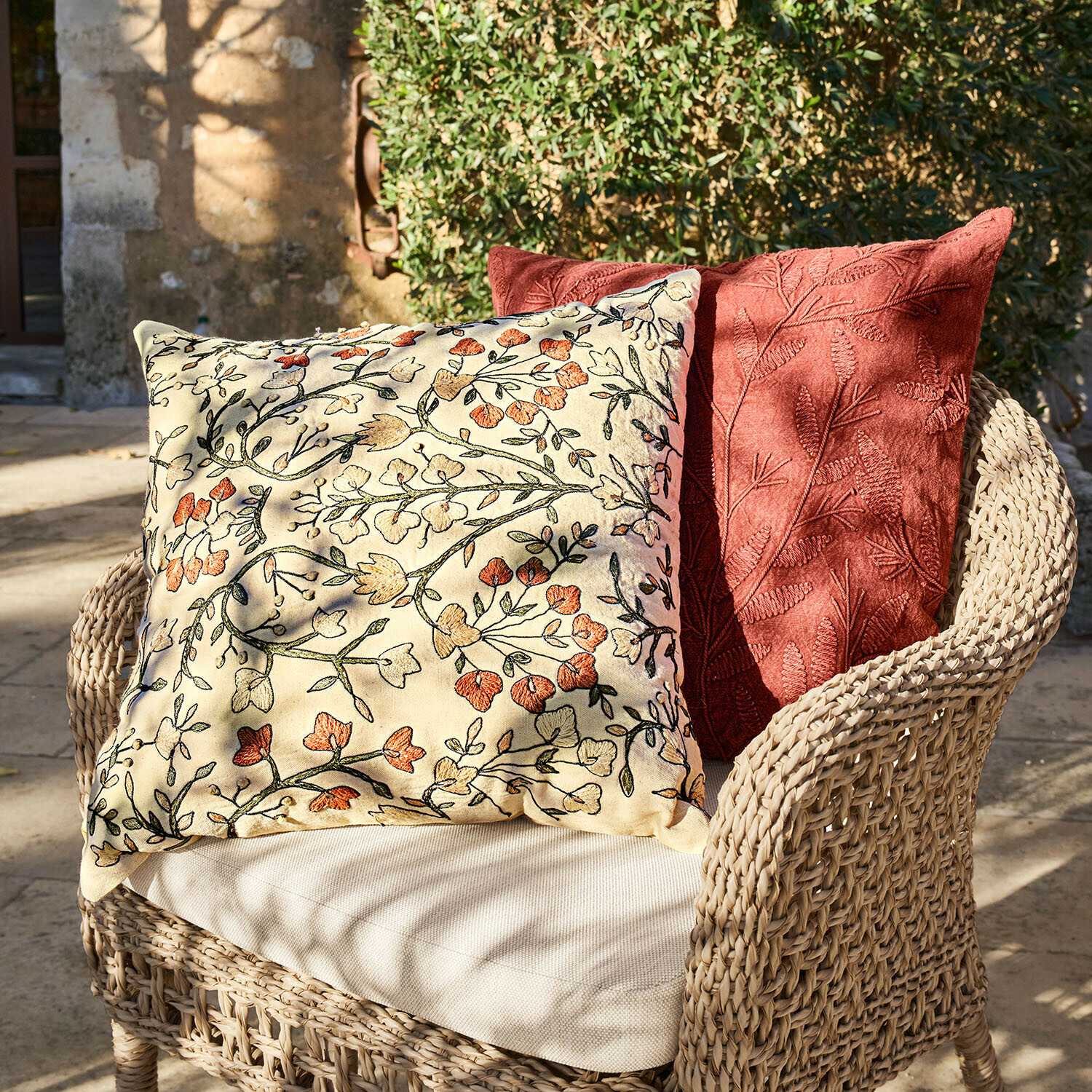 Set de 2 fundas de cojín Calévine Dos cojines decorativos con bordado floral y de hojas sobre sillón de mimbre en exterior
