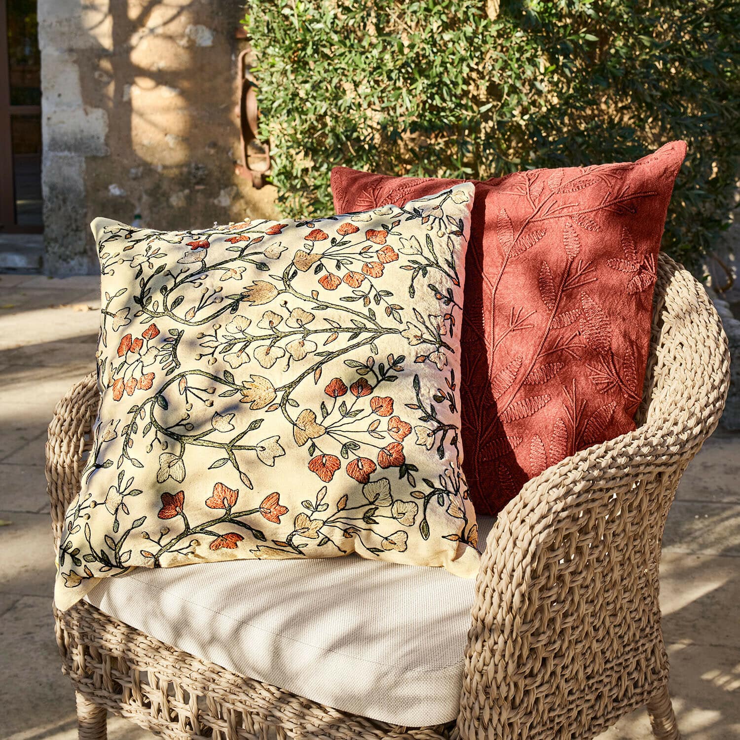 Set de 2 fundas de cojín Calévine Dos cojines decorativos con bordado floral y de hojas sobre sillón de mimbre en exterior
