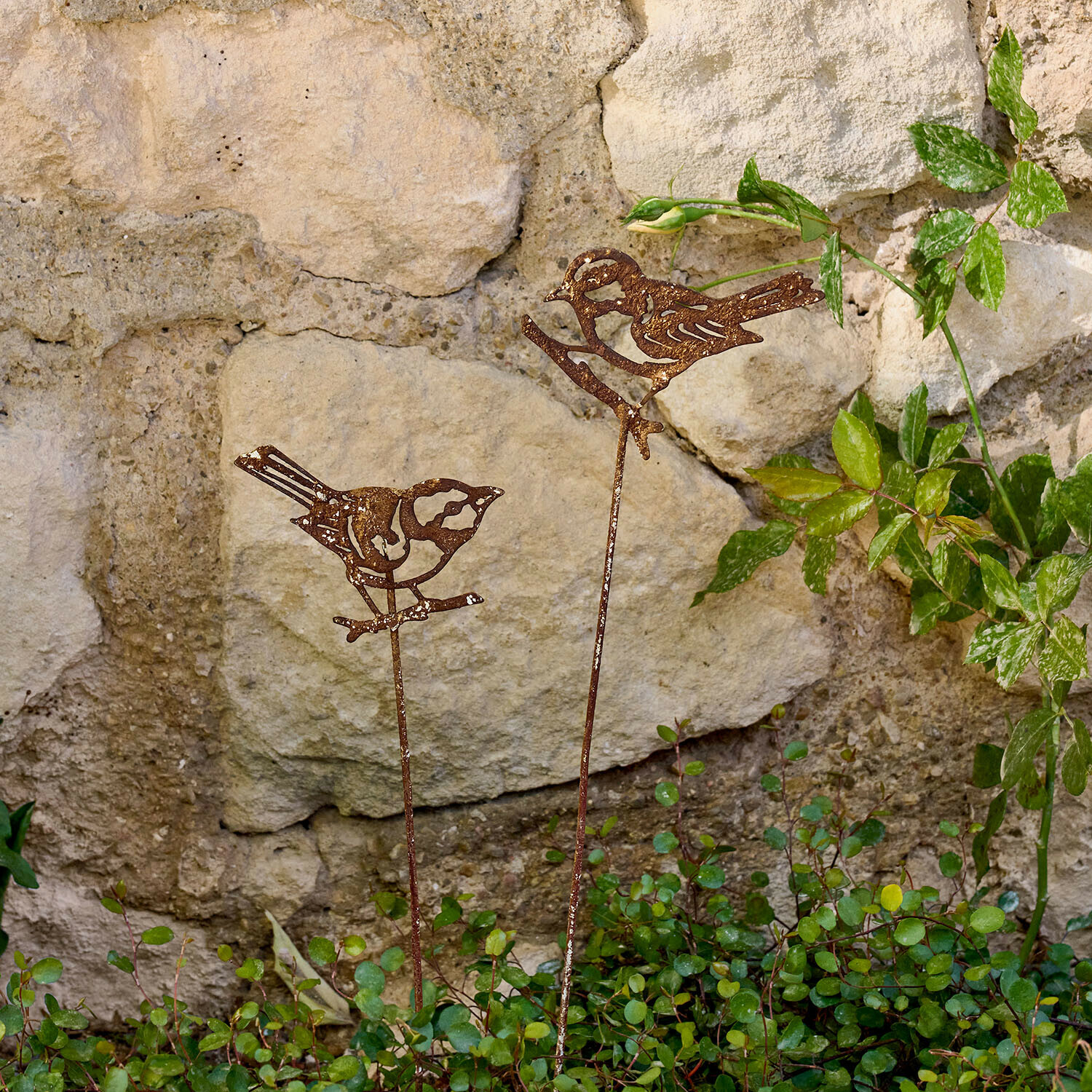 Set de 2 estacas de jardín Pelvaron Adornos de jardín de metal oxidado con forma de pájaros sobre varas frente a muro de piedra
