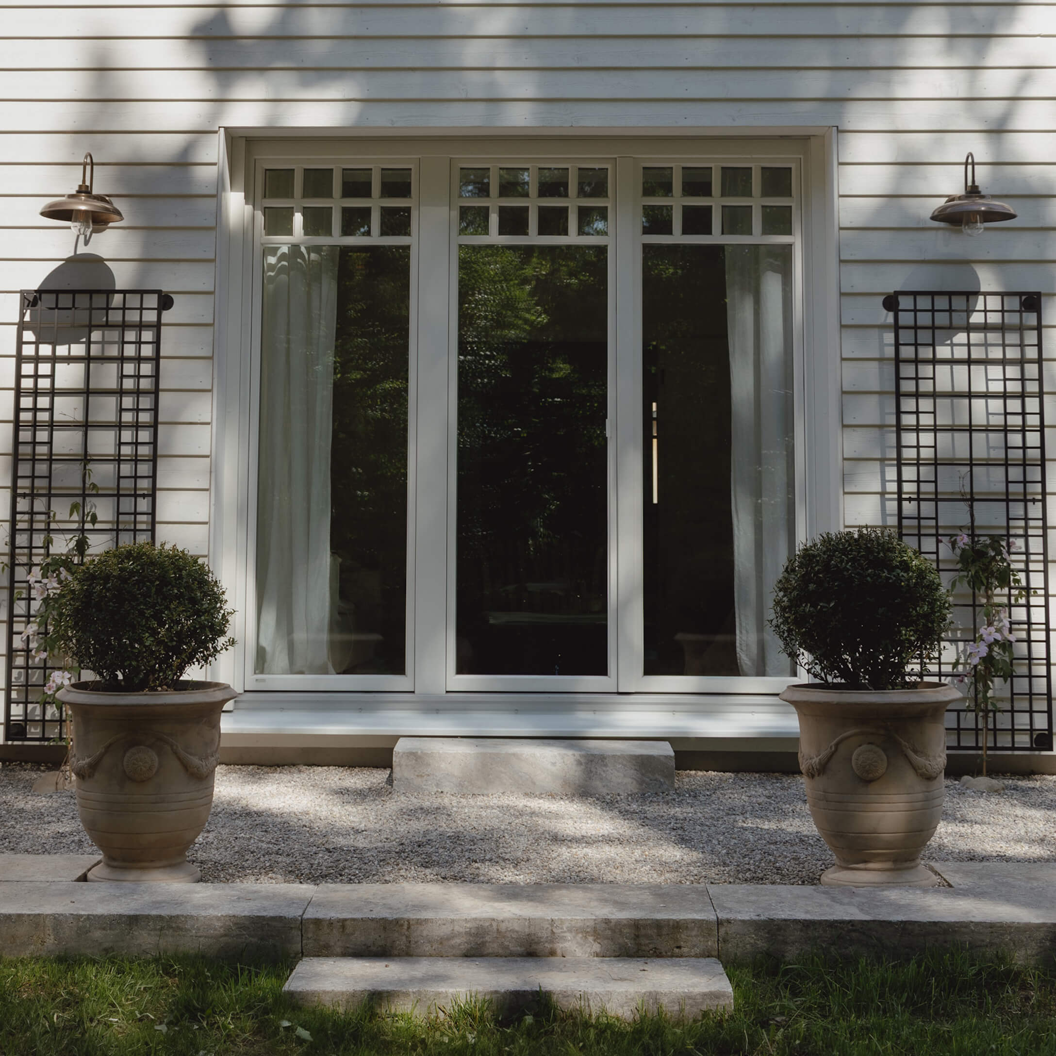Entrada de casa con puerta blanca y dos grandes macetas de piedra con plantas verdes sobre grava
