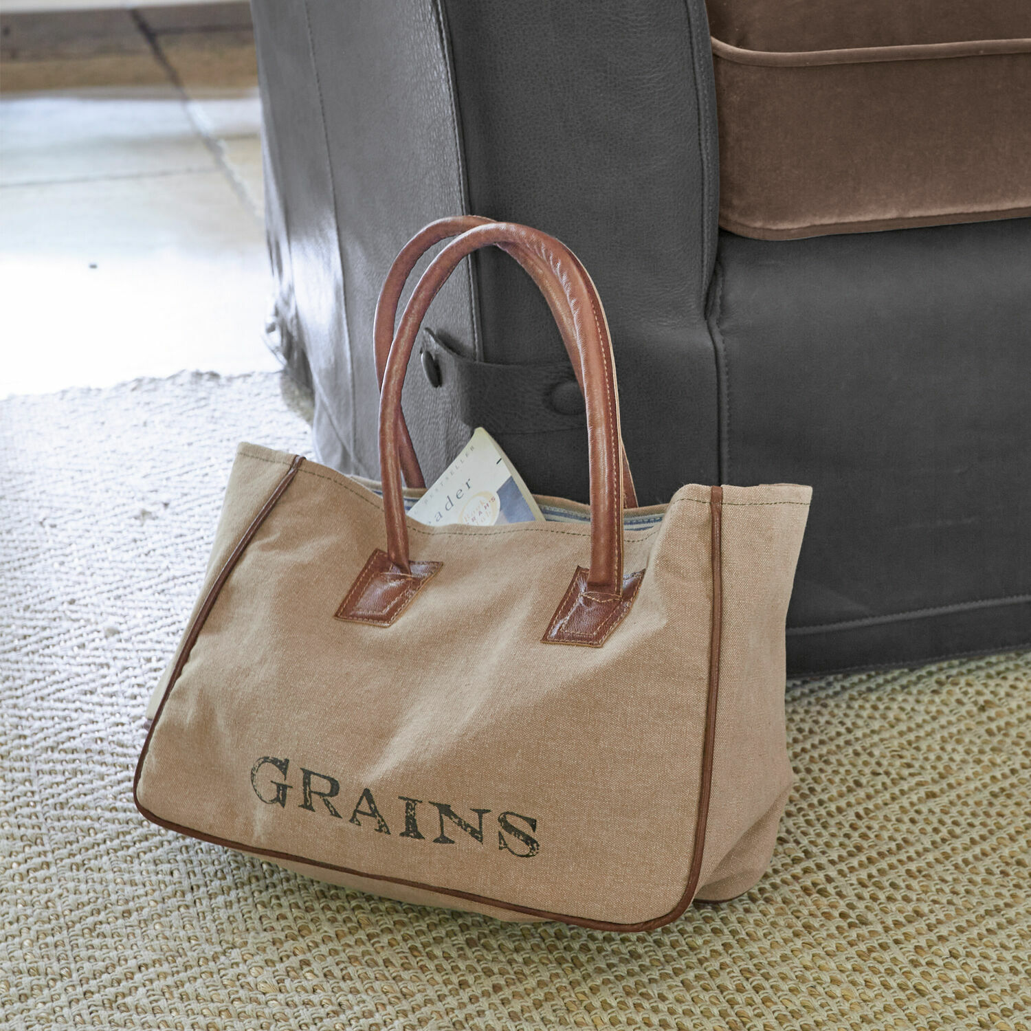 Bolsa beige de tela con asas de cuero marrón y texto GRAINS sobre alfombra junto a sofá