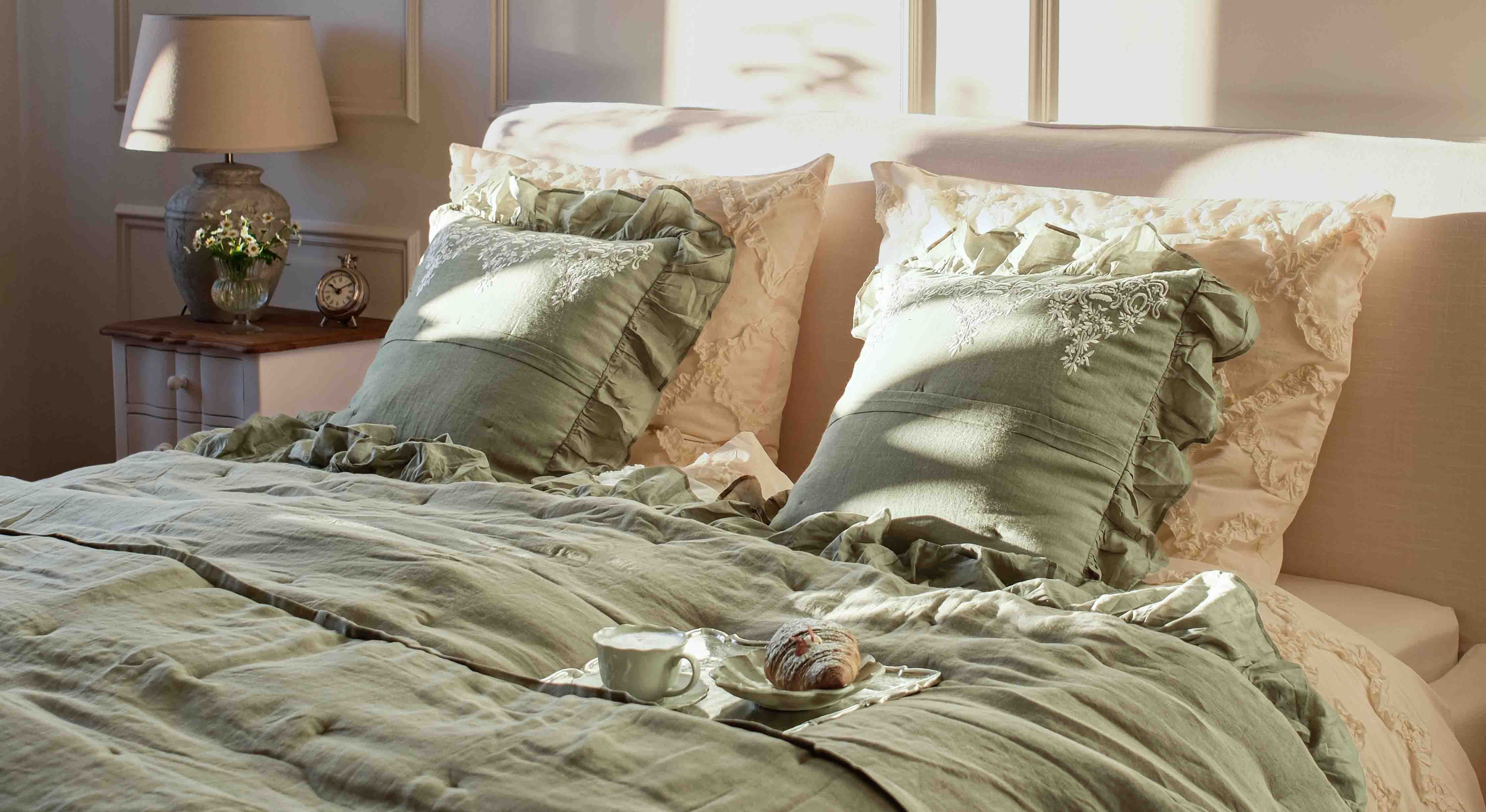 Ropa de cama verde de lino con cojines decorativos y fundas crema en dormitorio con lámpara y reloj