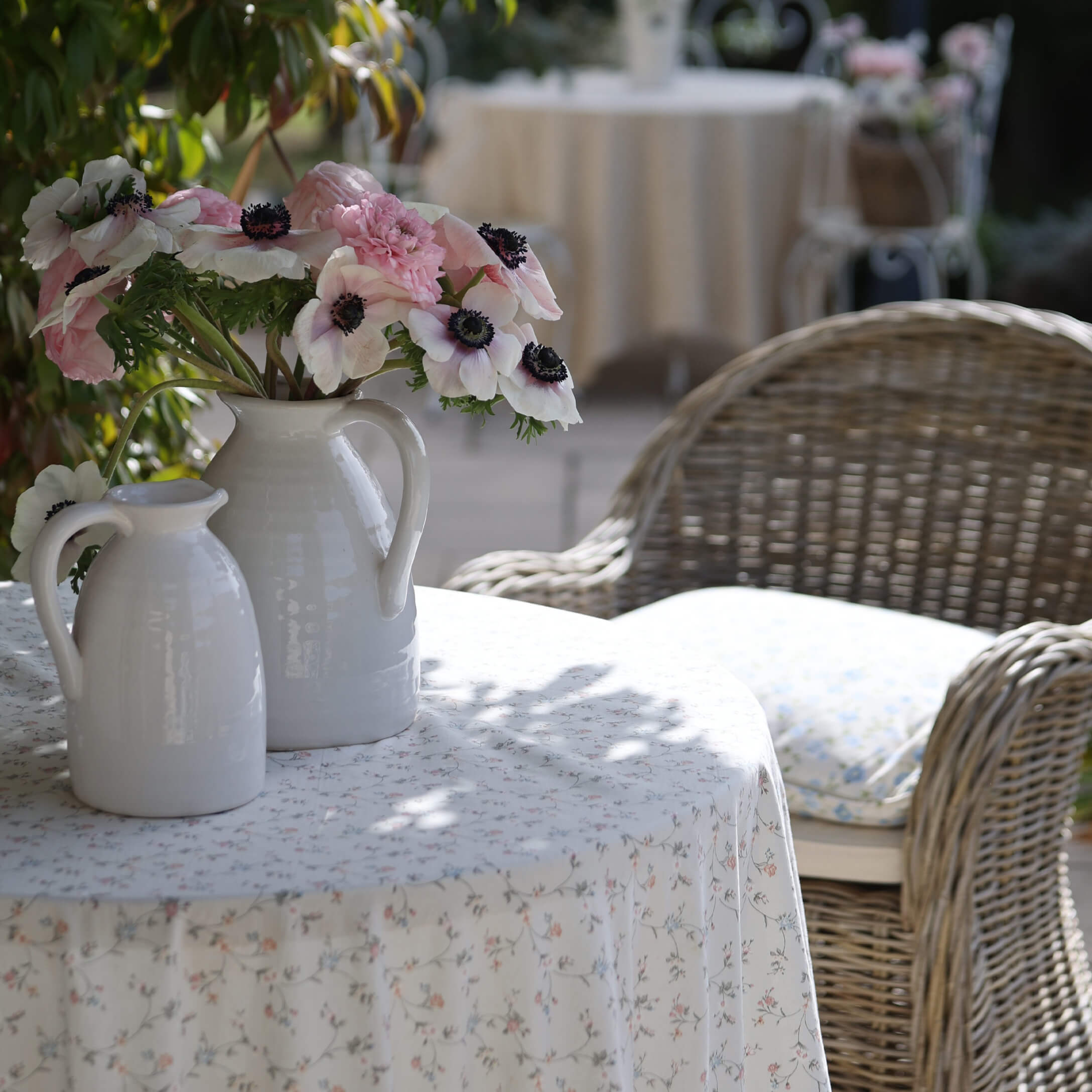 Jarras de cerámica blanca con flores rosas y blancas sobre mesa con mantel floral en jardín