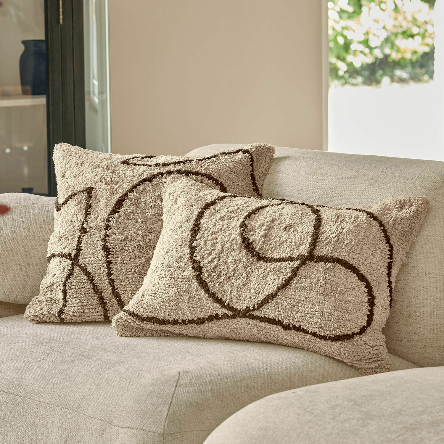Cojines decorativos beige de material suave con diseño geométrico marrón sobre sofá claro