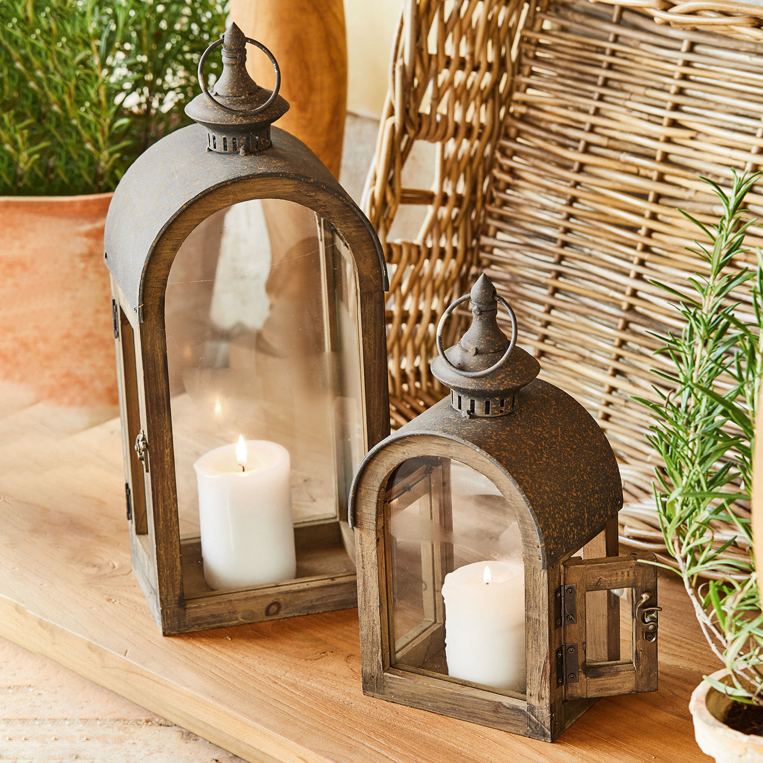 Set de 2 faroles Jadon Faroles de madera con tapa metálica oxidada y velas blancas sobre mesa de madera, decoración rústica