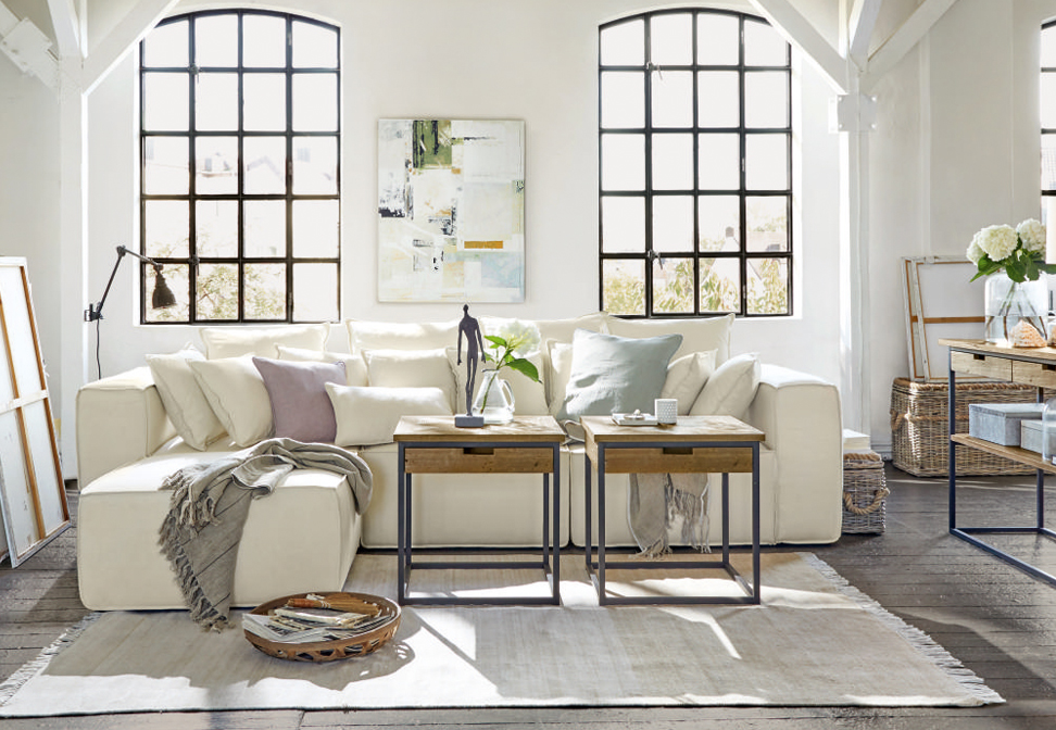 Sofá esquinero blanco con cojines y mesas de madera en sala luminosa con grandes ventanas