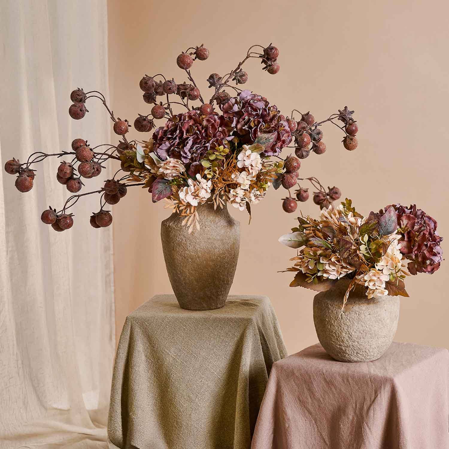 Dos ramos decorativos de flores secas en jarrones beige sobre mesas cubiertas con tela