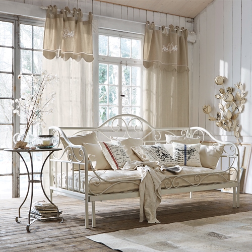 Sofá metálico blanco con cojines beige y cortinas claras en salón rústico luminoso