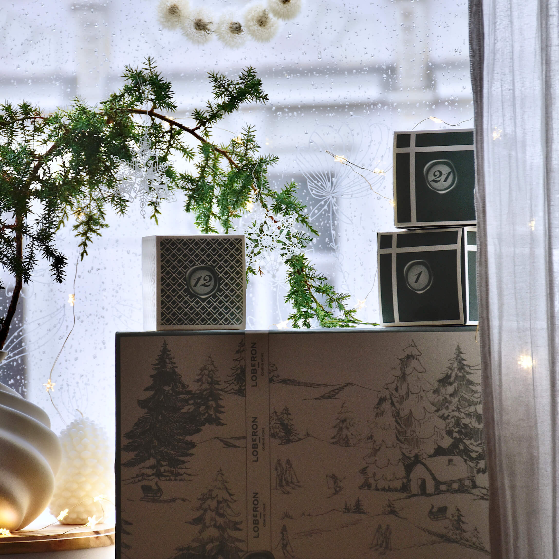 Cajas decorativas con motivo invernal y rama verde con copos blancos frente a ventana con lluvia