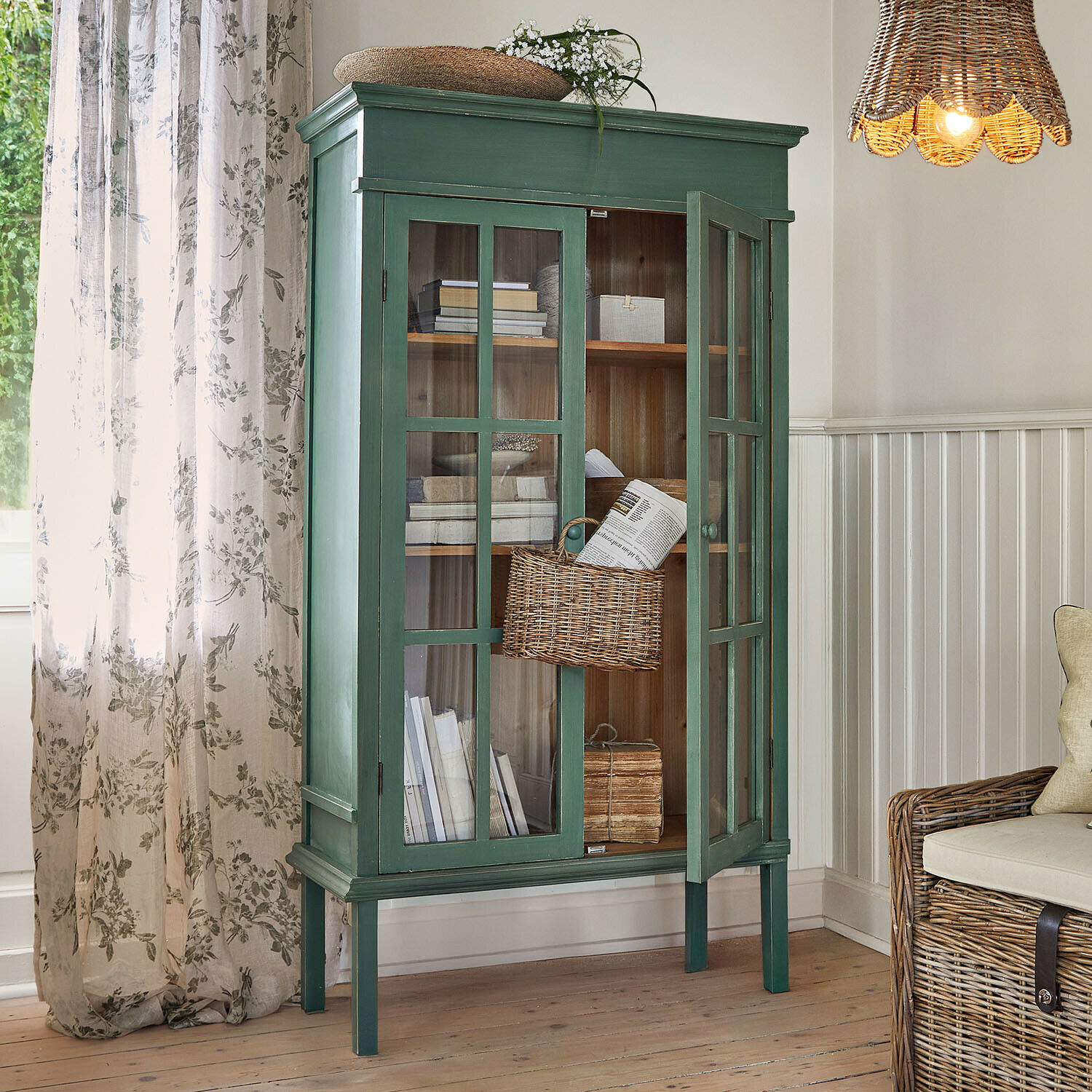Vitrina Faymere Vitrina de madera verde con puertas de cristal y cesta de mimbre en salón con luz natural