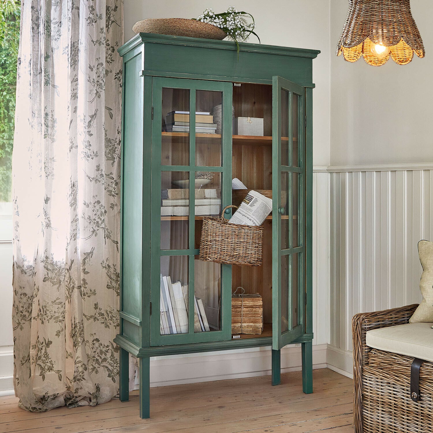 Vitrina Faymere Vitrina de madera verde con puertas de cristal y cesta de mimbre en salón con luz natural