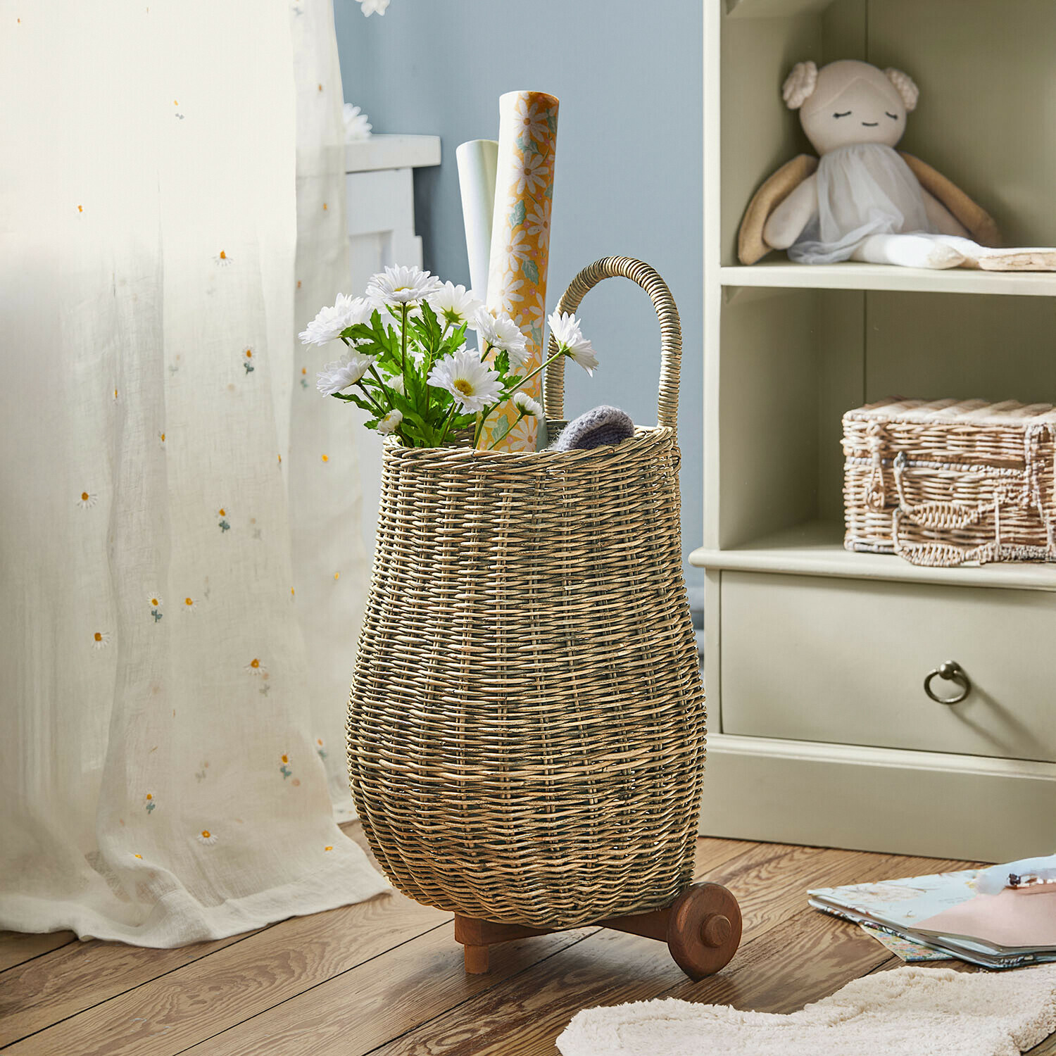 Cesta de mimbre natural con ruedas y asa, llena de flores blancas y papeles decorativos