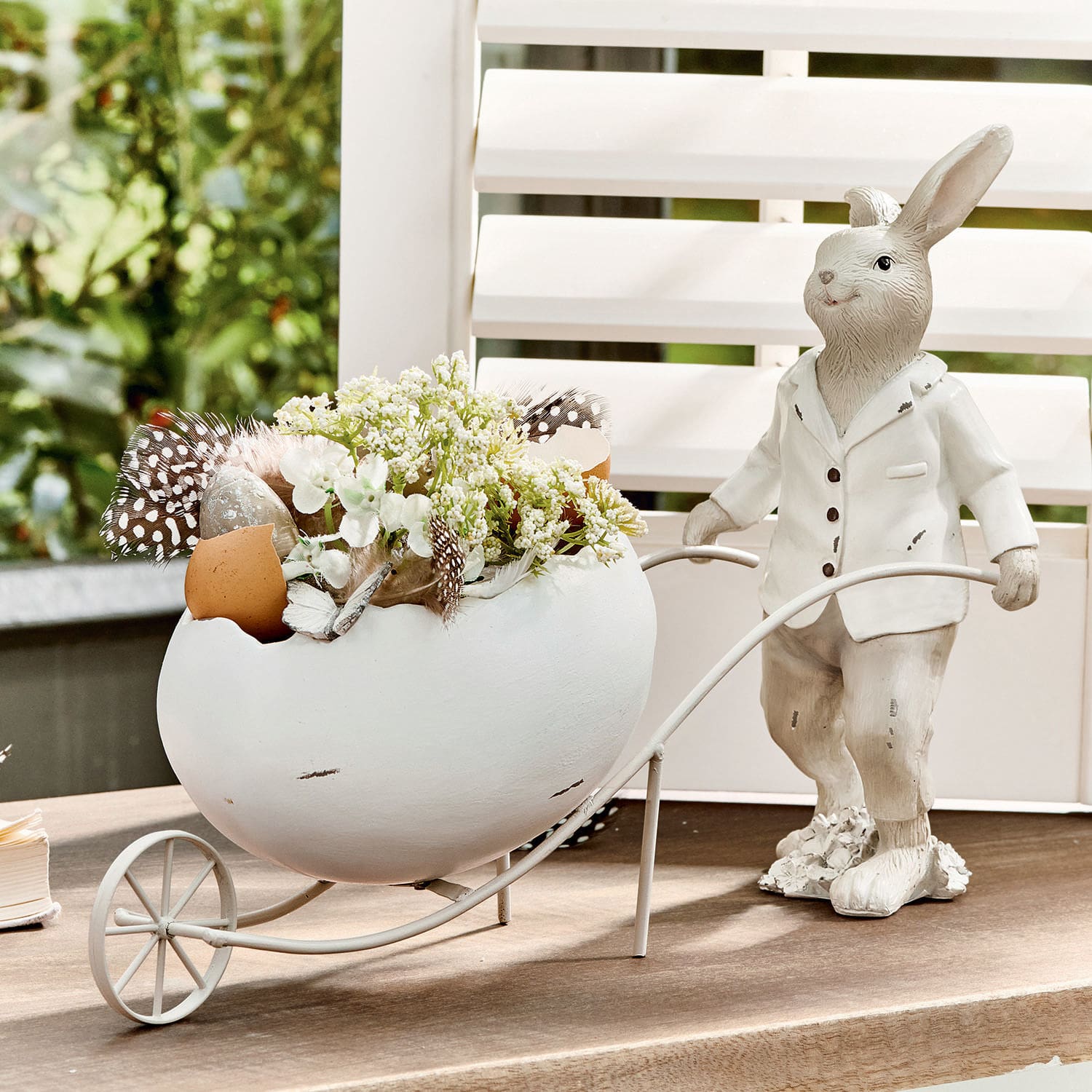 Conejo Jaris Conejo decorativo blanco con huevo de cerámica y flores en carrito metálico sobre mesa.
