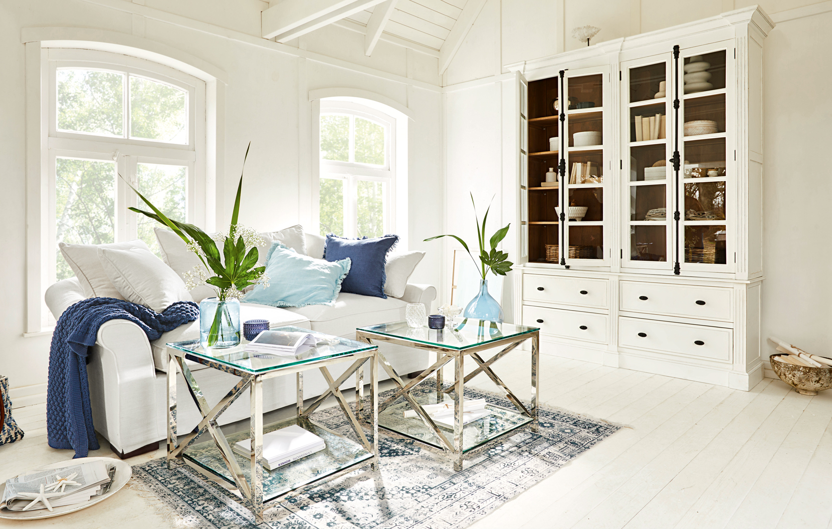 Sala luminosa con sofá blanco, dos mesas de cristal y vitrina blanca frente a grandes ventanas